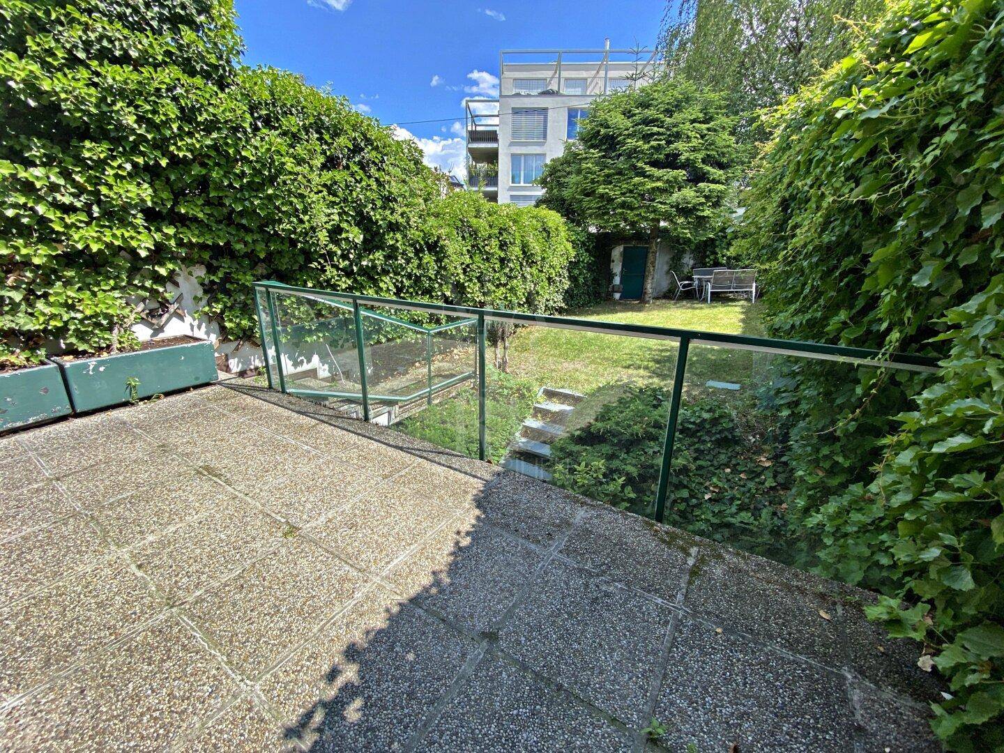 Außenbereich mit gepflasterter Terrasse, Glasgeländer und Treppe zum unteren Gartenbereich.