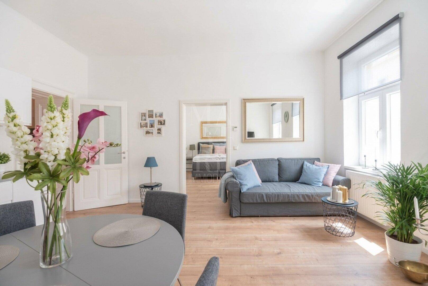 Heller Wohn- und Essbereich mit Holzboden, Sofa und Blick in ein angrenzendes Schlafzimmer.