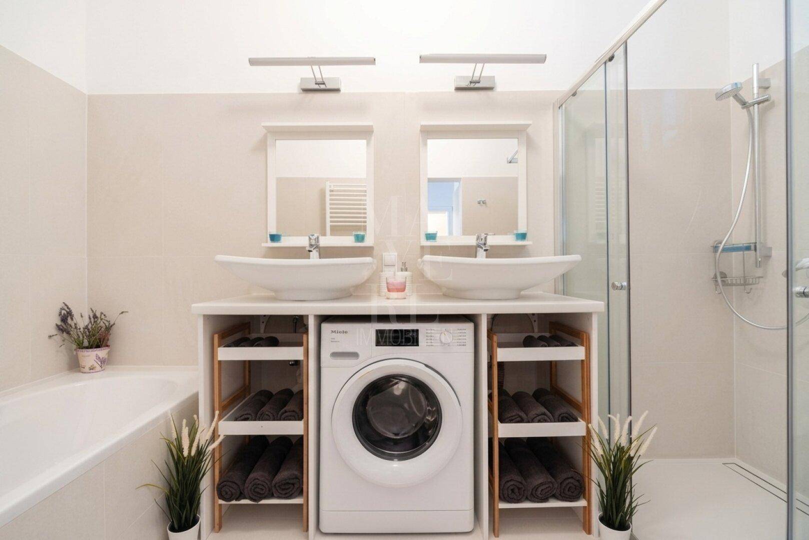 Helles Badezimmer mit Doppelwaschbecken, großer Dusche und integrierter Waschmaschine.