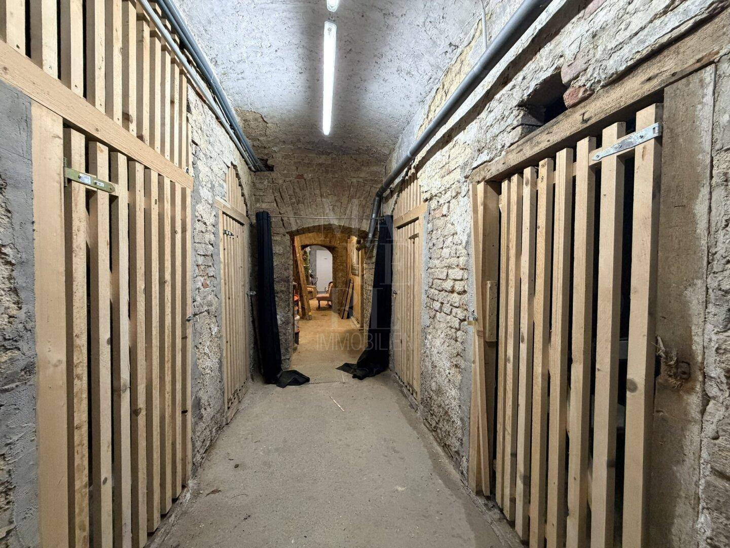 Langer Kellergang mit gemauerten Wänden und Holzverschlägen für Lagerzwecke.