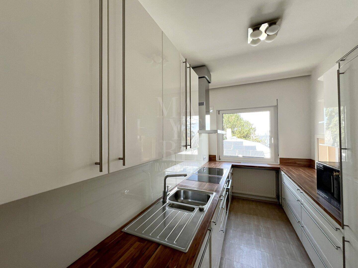 Moderne Küche mit weißen Hochglanzfronten, Holzarbeitsplatten und Fenster mit Ausblick.