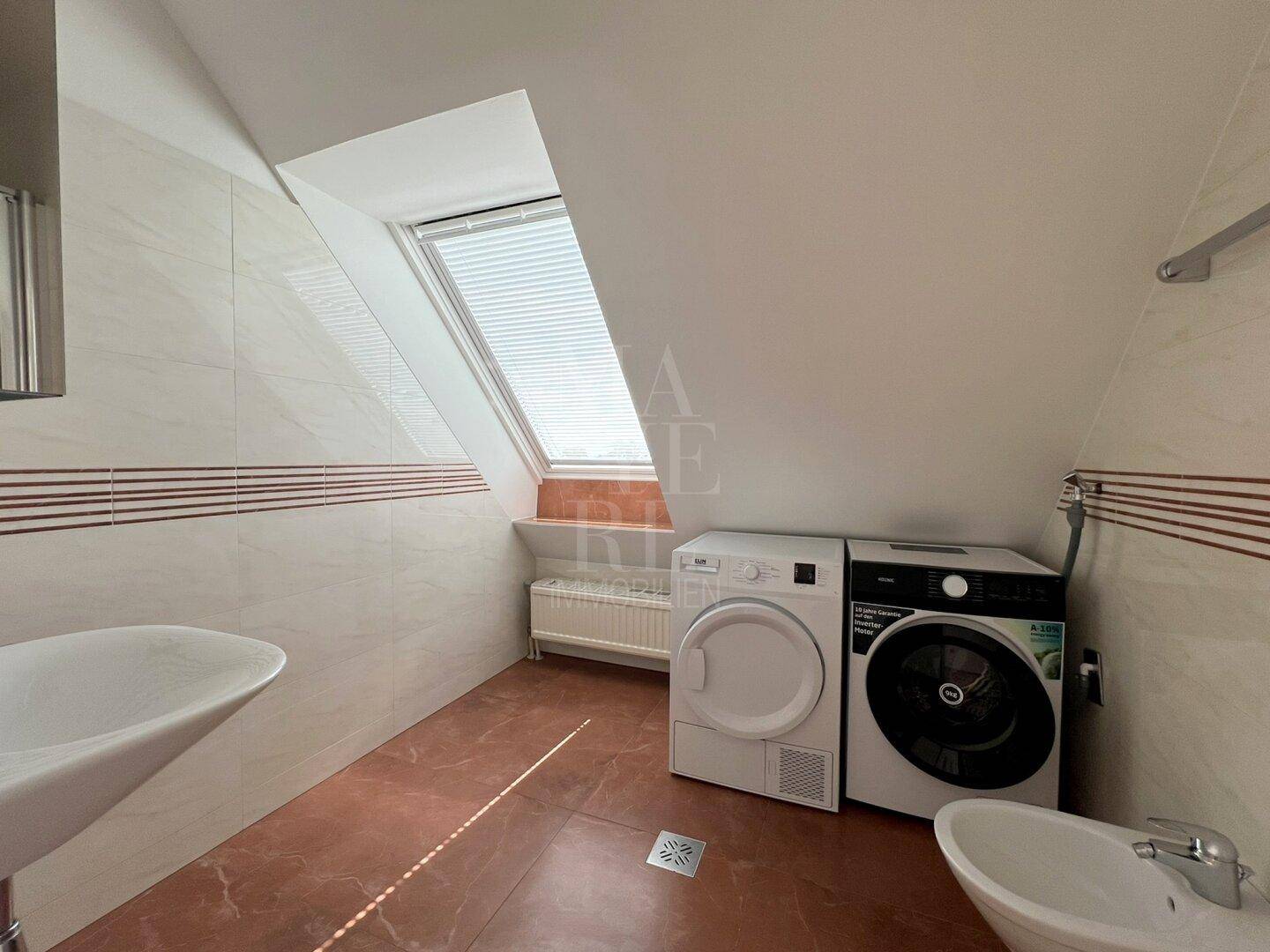 Badezimmer mit Dachfenster, Waschmaschine, Trockner und Fliesenboden in warmen Farbtönen.