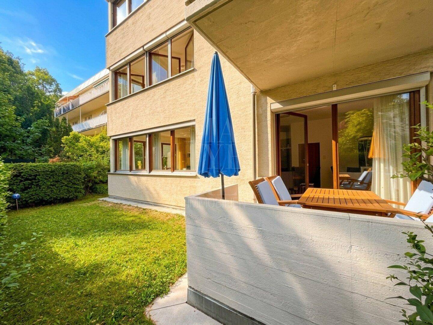 Terrasse mit blauem Sonnenschirm und Blick auf die grüne Umgebung.