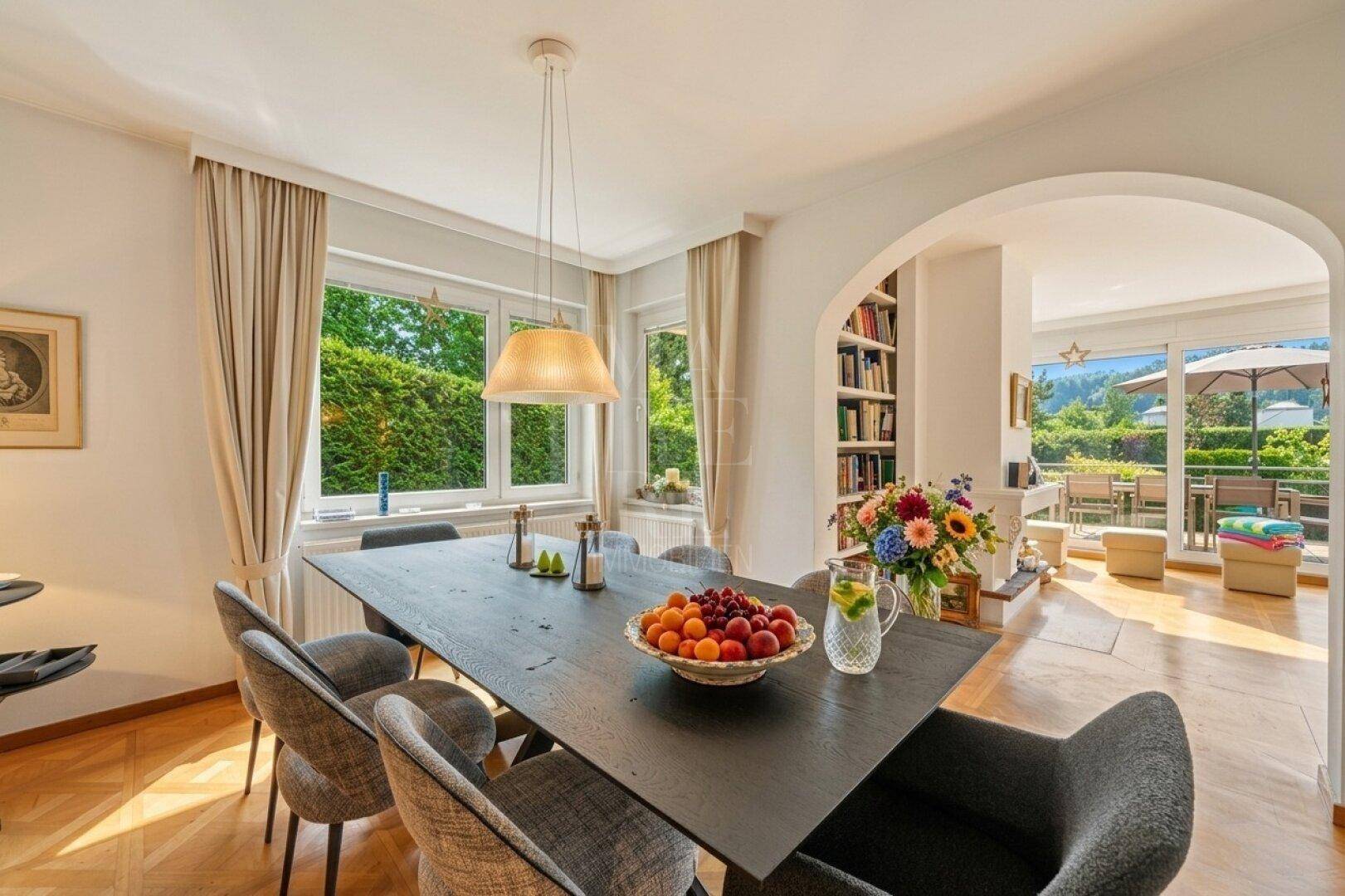 Eleganter Essbereich mit großem Holztisch, modernen Stühlen und Blick ins Grüne durch die Fenster.