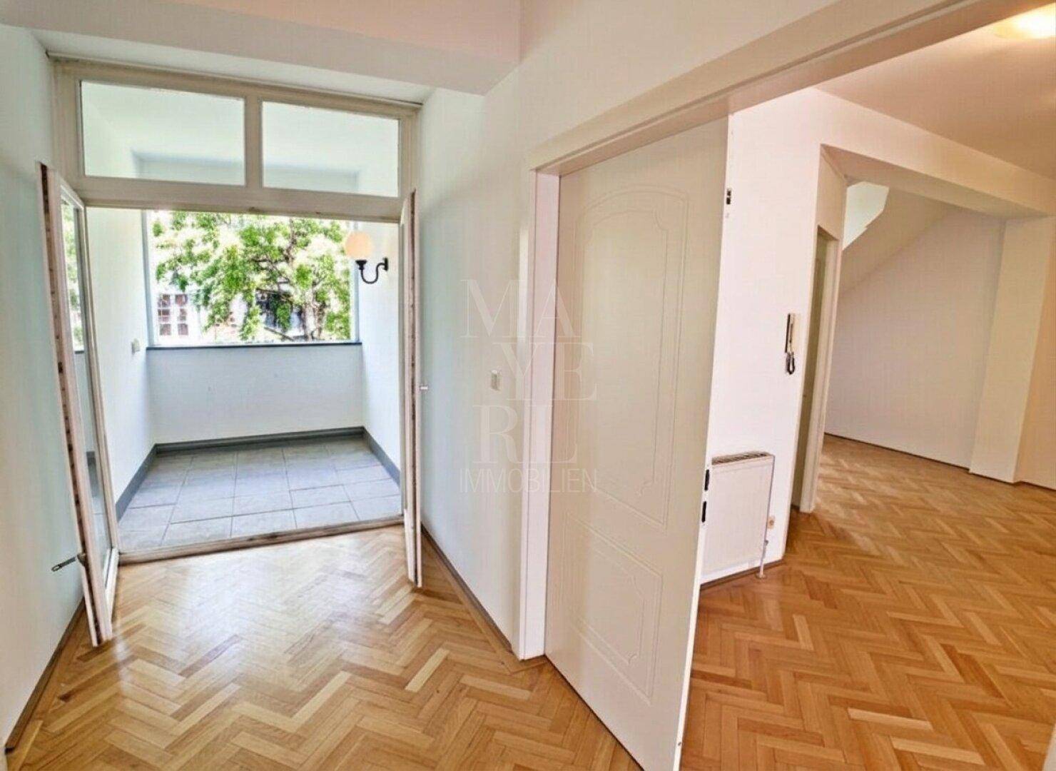Flur mit Fischgrätparkett und Zugang zu einem kleinen, gefliesten Balkon mit Blick ins Grüne.
