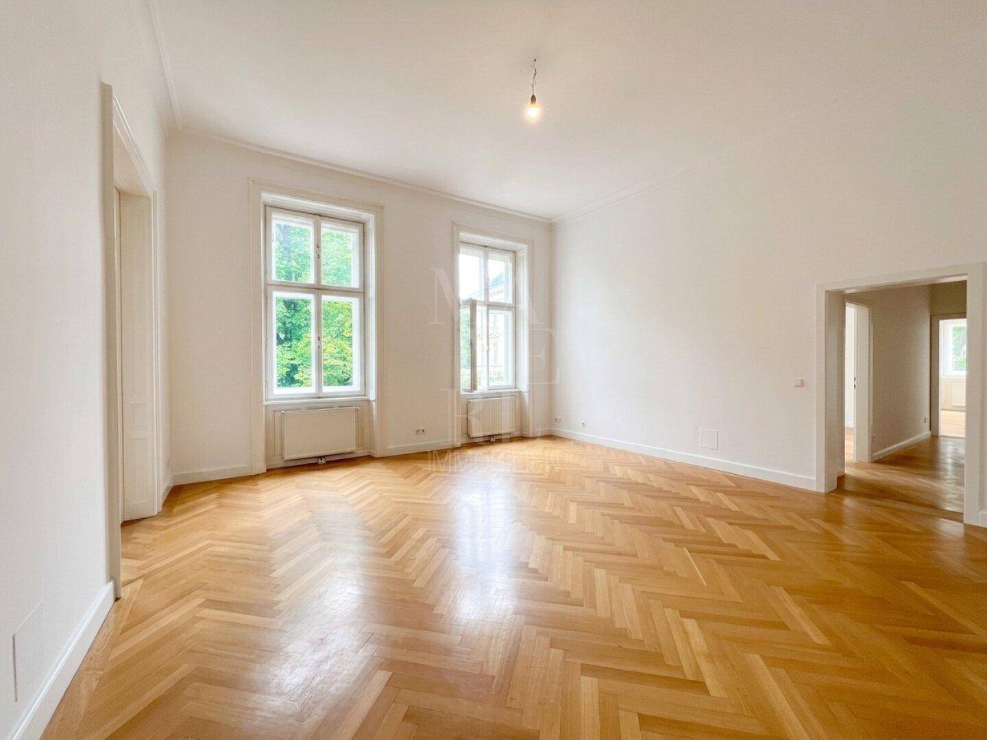 Geräumiges Wohnzimmer mit klassischem Fischgrätparkett und viel Tageslicht durch hohe Fenster.