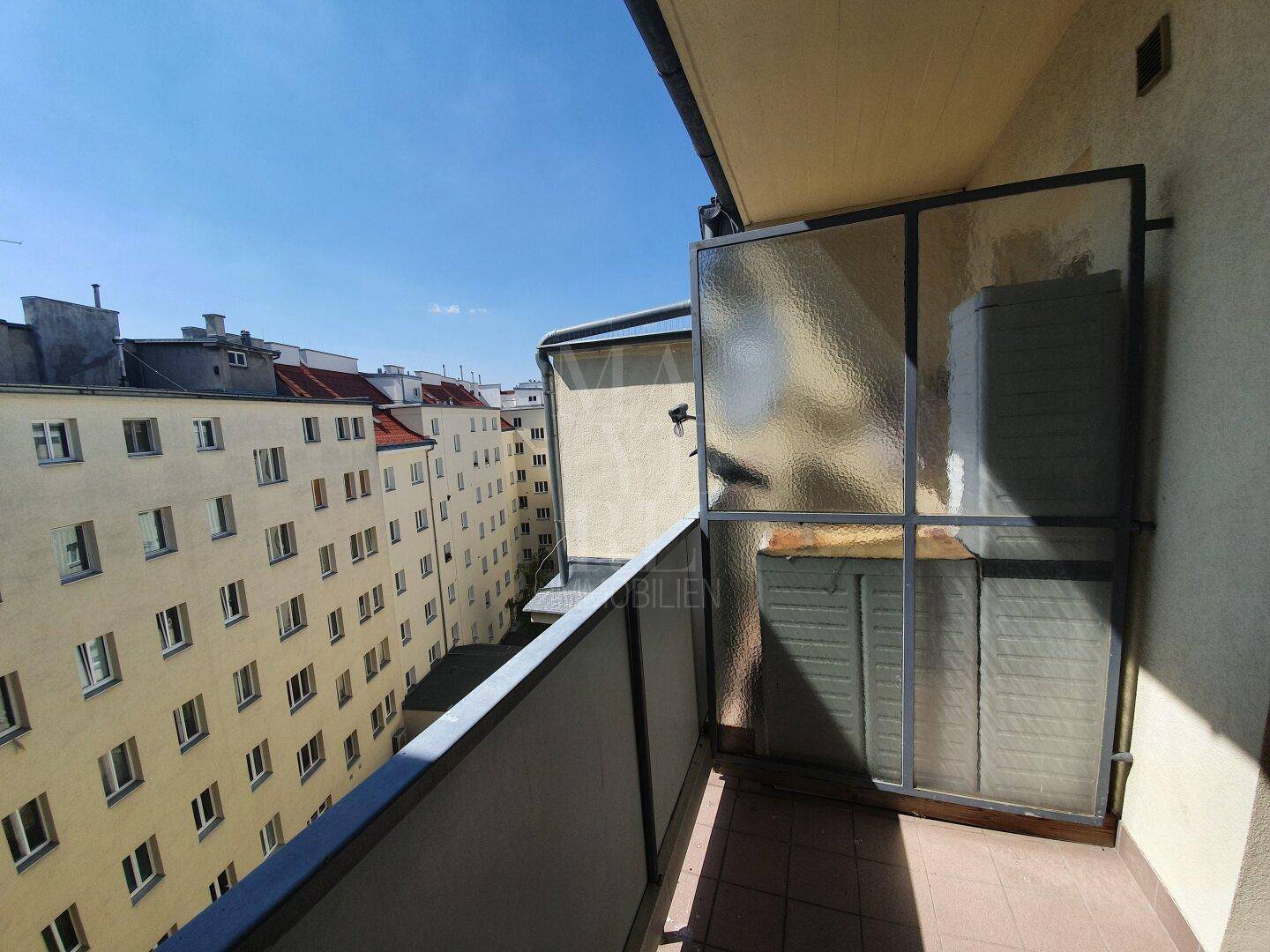 Balkon mit Blick auf die umliegenden Wohngebäude und sonnigem Wetter, mit Glasabtrennung.