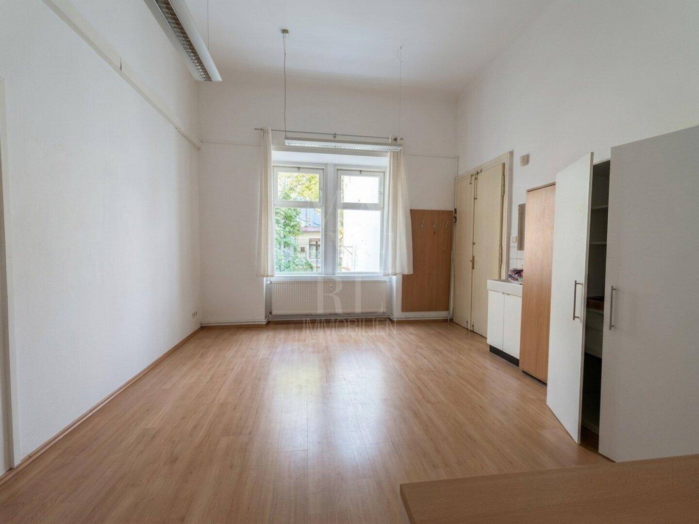 Ein heller Raum mit Fenster, Holzboden und einfachen weißen Schränken an der Wand.
