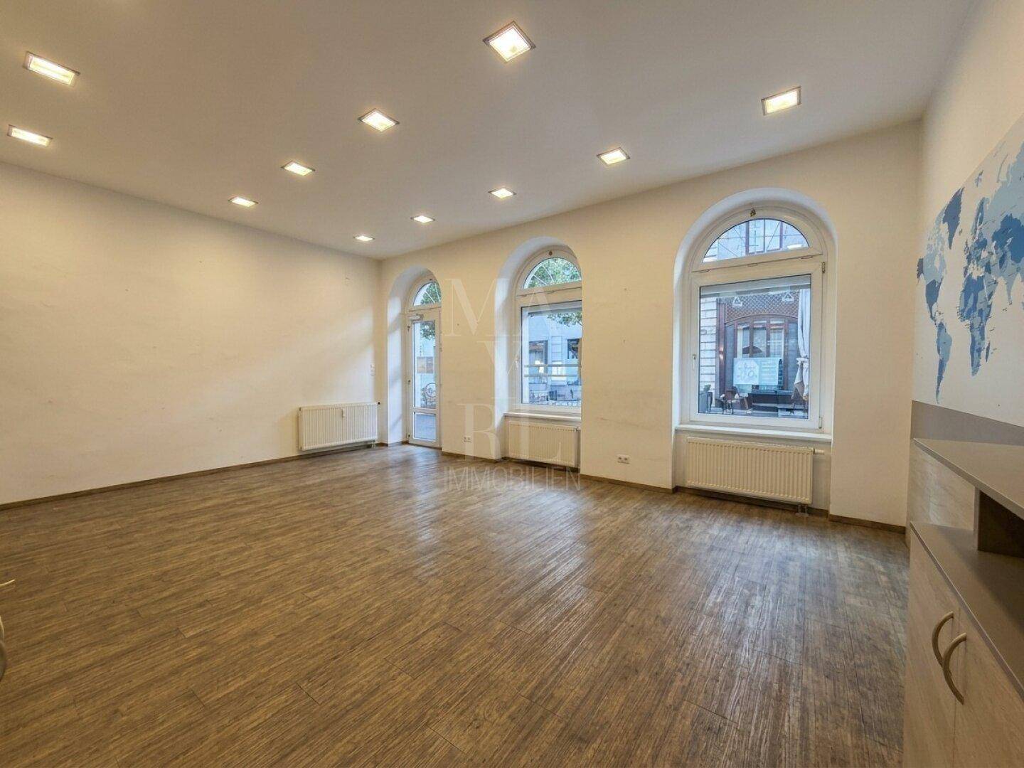 Ein großer, leerer Raum mit Holzboden, Einbaustrahlern und drei hohen Bogenfenstern zur Straße.
