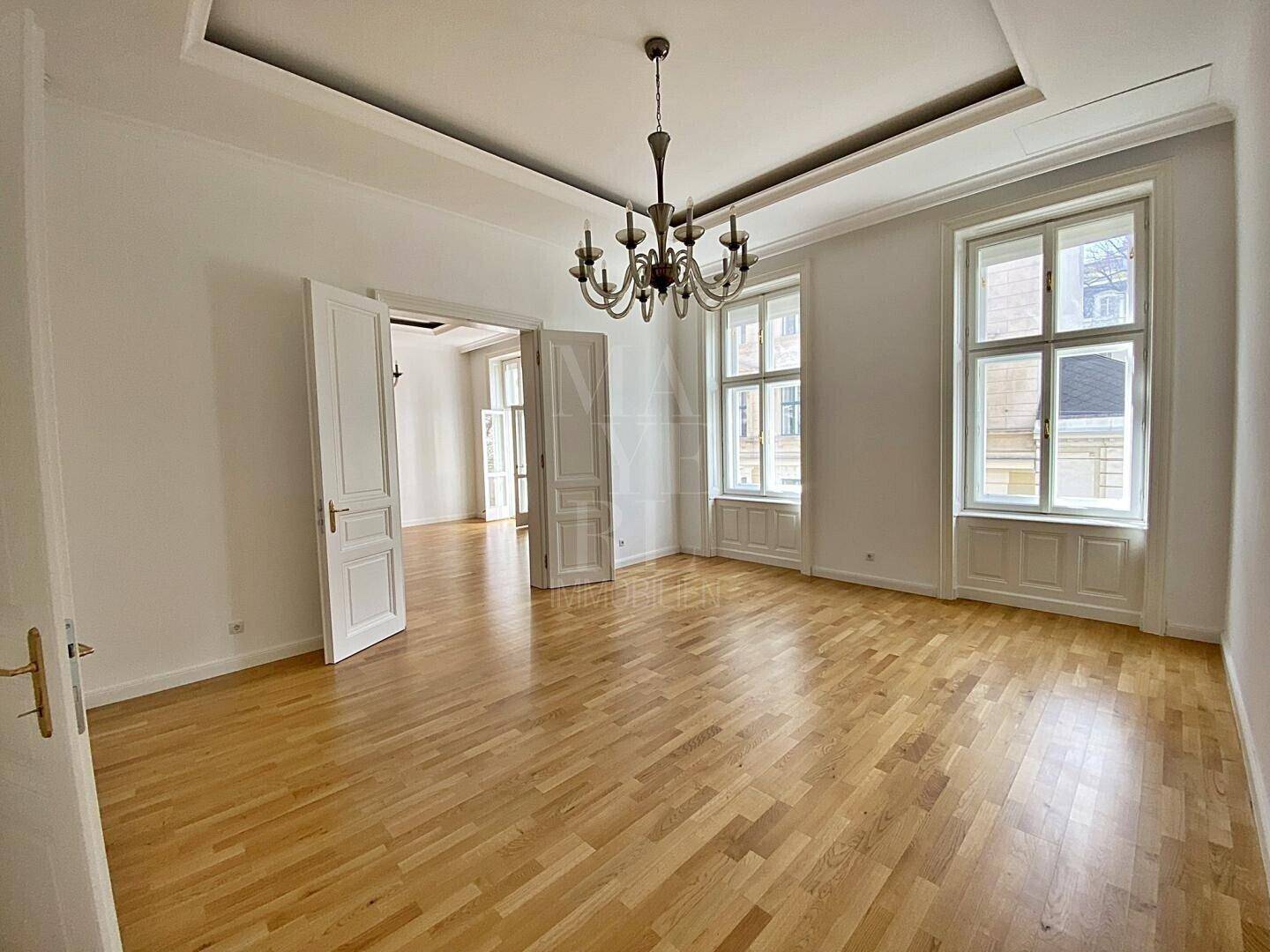 Geräumiges Wohnzimmer mit klassischem Kronleuchter, Parkettboden und hohen Fenstern.