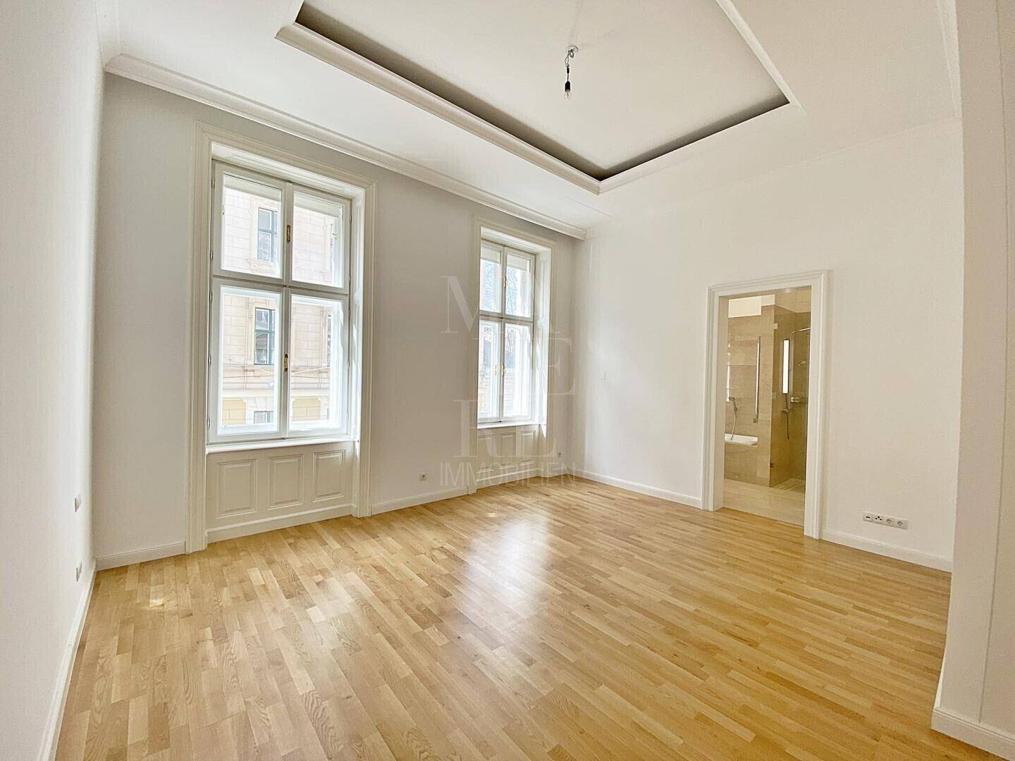 Heller Raum mit Parkettboden, hohen Decken und Blick in ein angrenzendes Badezimmer.
