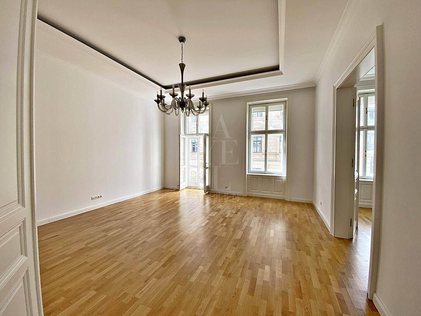 Heller Raum mit Parkettboden, hohen Decken und Zugang zu einem französischen Balkon.
