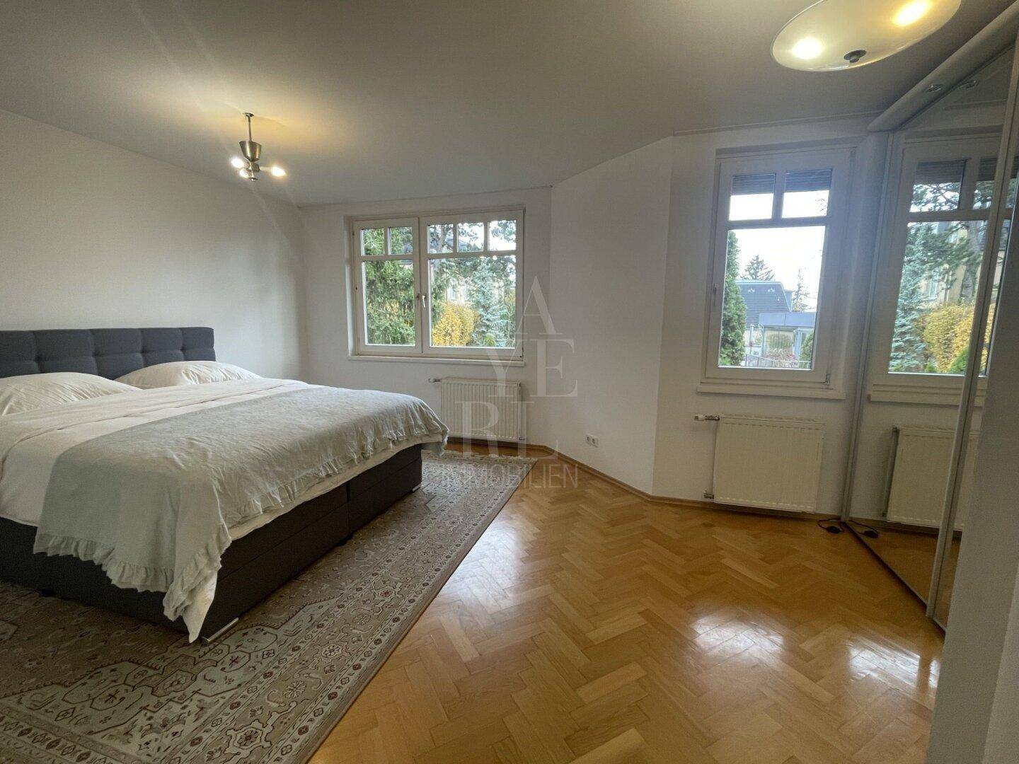 Helles Schlafzimmer mit Parkettboden, großem Bett und mehreren Fenstern mit Grünblick.