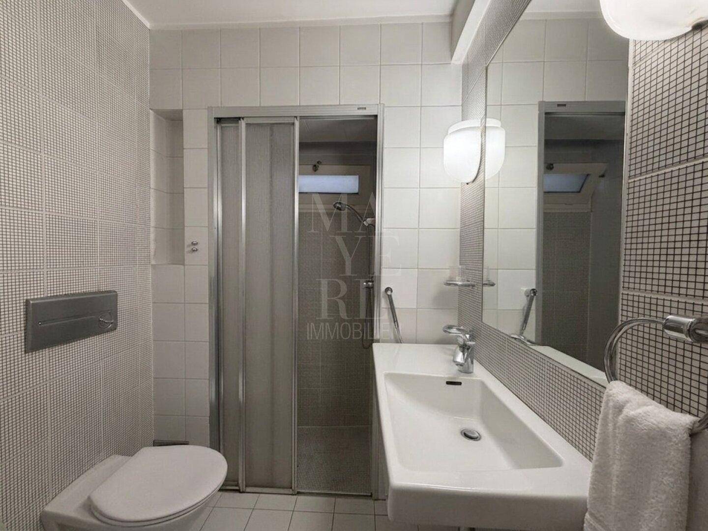 Helles Badezimmer mit weißen Fliesen, Dusche und modernem Waschbecken.
