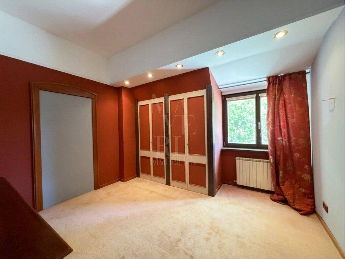 Schlafzimmer mit roter Wandfarbe, Teppichboden und Fenster mit Blick ins Grüne.