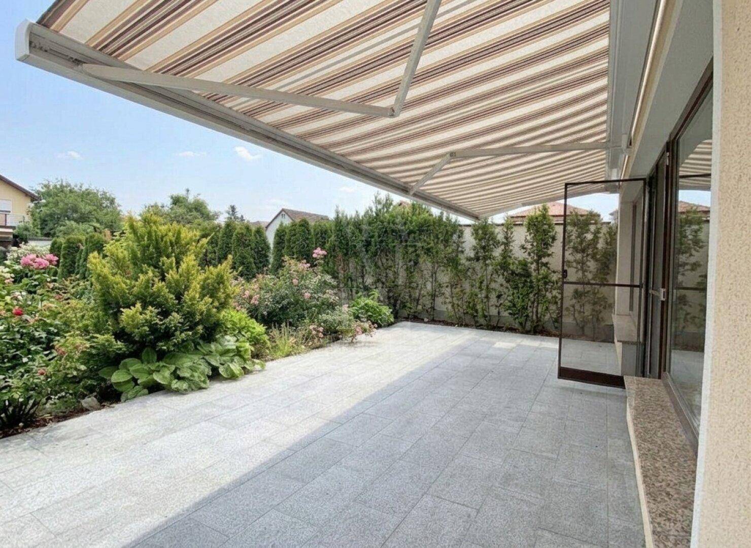 Große geflieste Terrasse mit Markise, umgeben von üppiger Bepflanzung und Blumen.