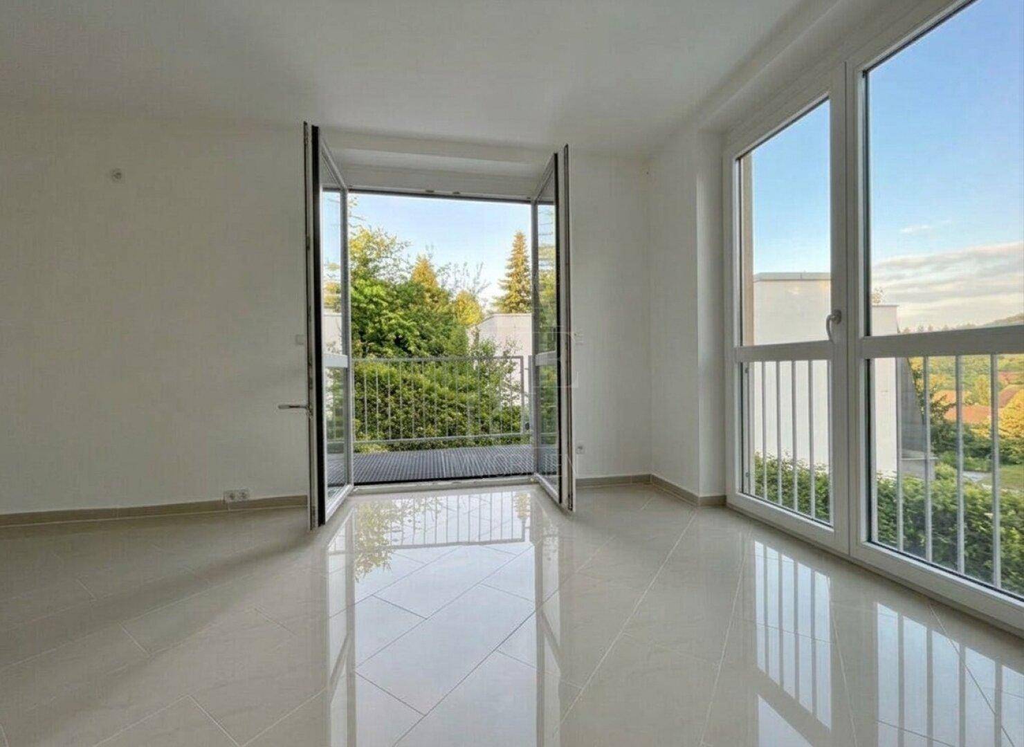 Helles Zimmer mit großen Fenstern, Fliesenboden und Zugang zu einem französischen Balkon.