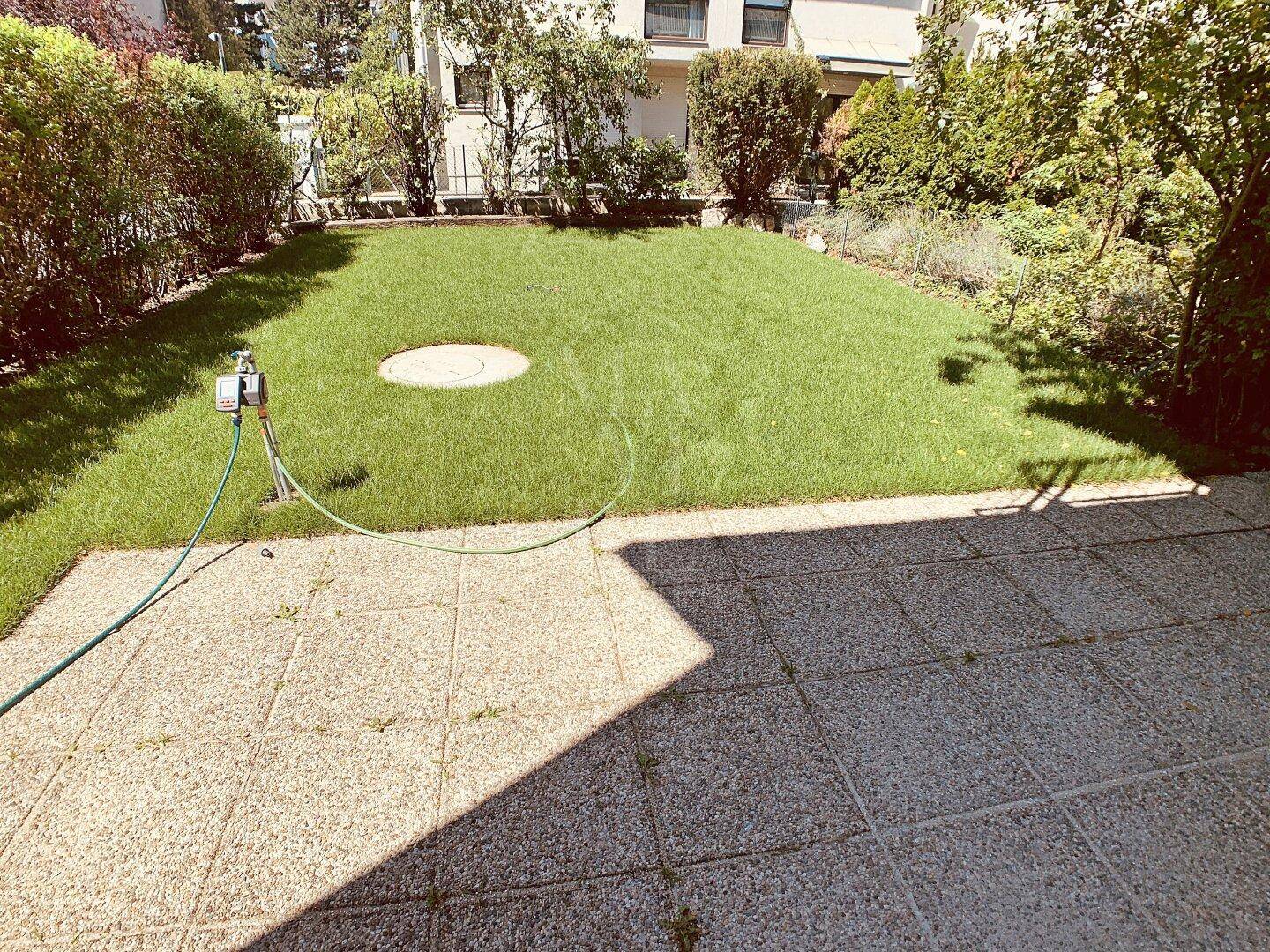 Gepflegter Gartenbereich mit Rasenfläche, Bewässerungssystem und einer gefliesten Terrasse.