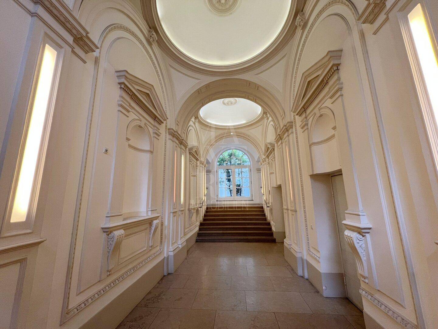 Prunkvoller Flur mit Stuckverzierungen, gewölbter Decke und einer eleganten Treppe am Ende.