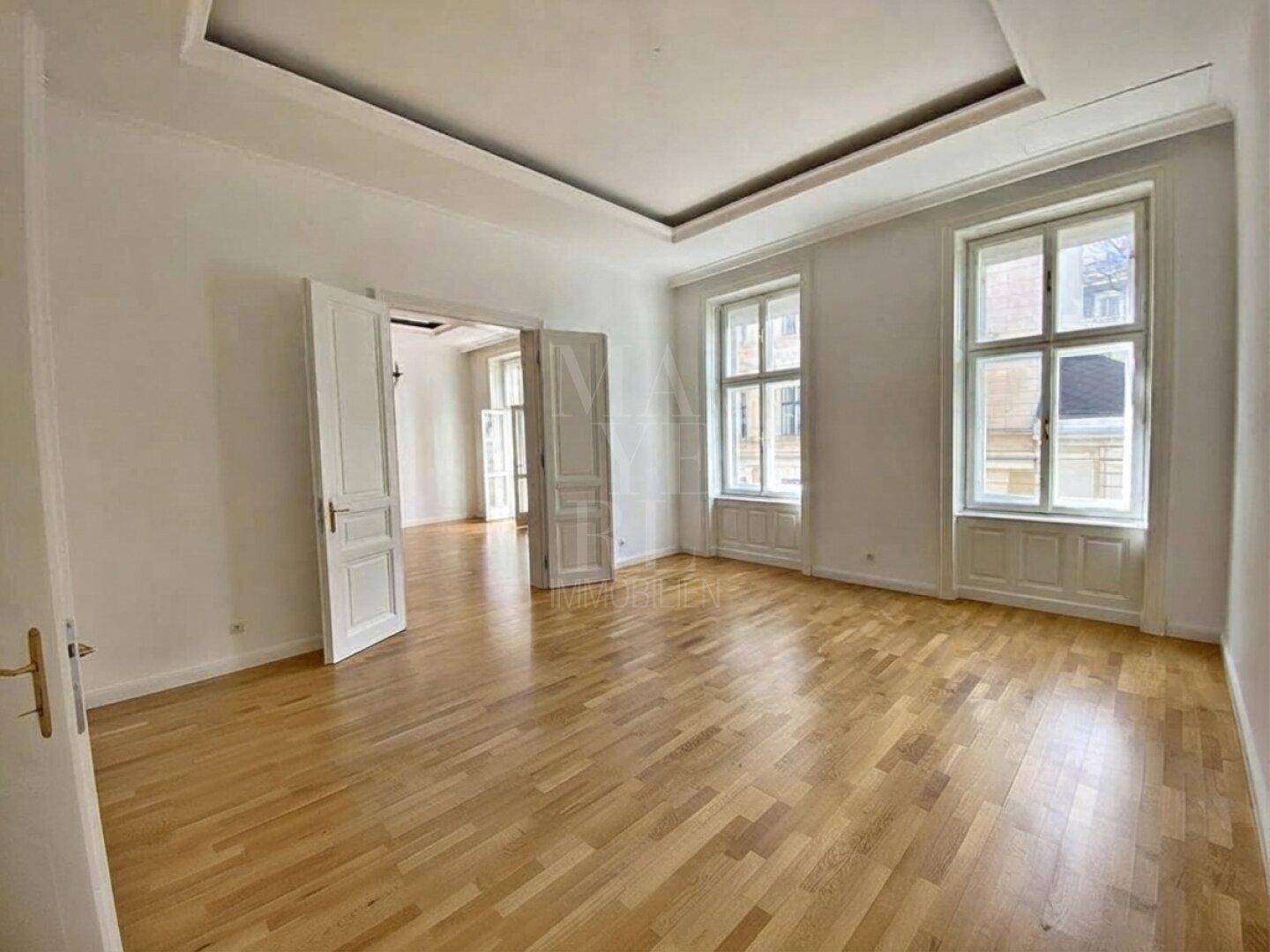 Geräumiges Wohnzimmer mit hellem Parkettboden, hohen Fenstern und mehreren Türöffnungen zu angrenzenden Räumen.