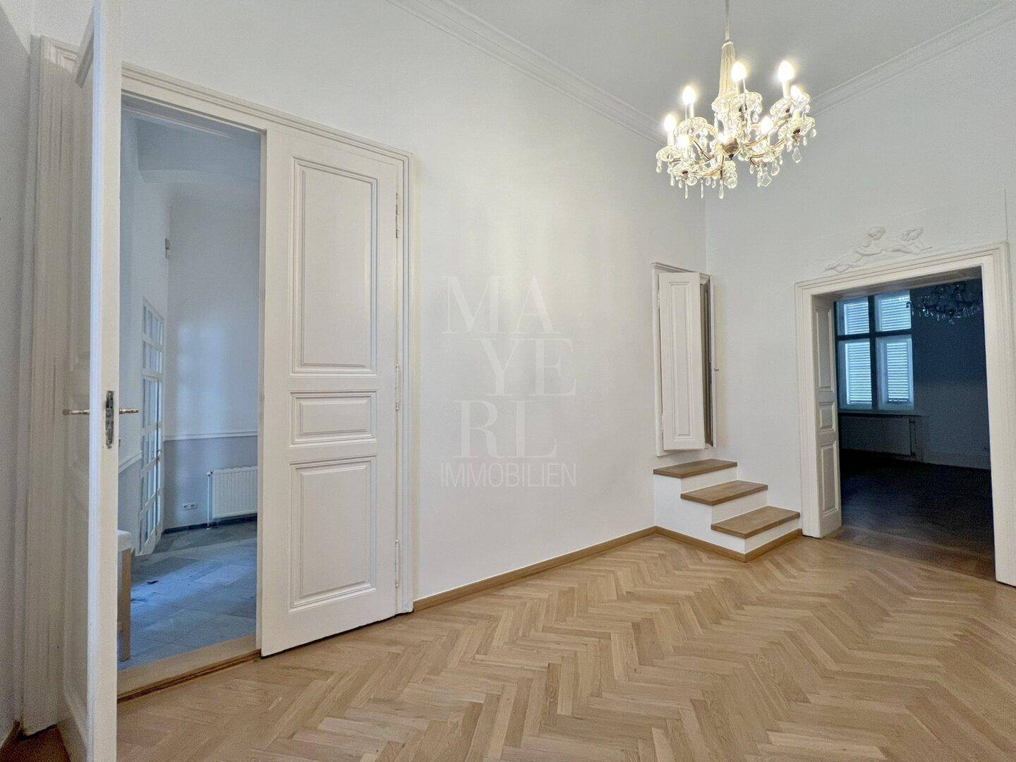 Eleganter Flur mit Fischgrätparkett, Kronleuchter und Zugang zu verschiedenen stilvollen Räumen.