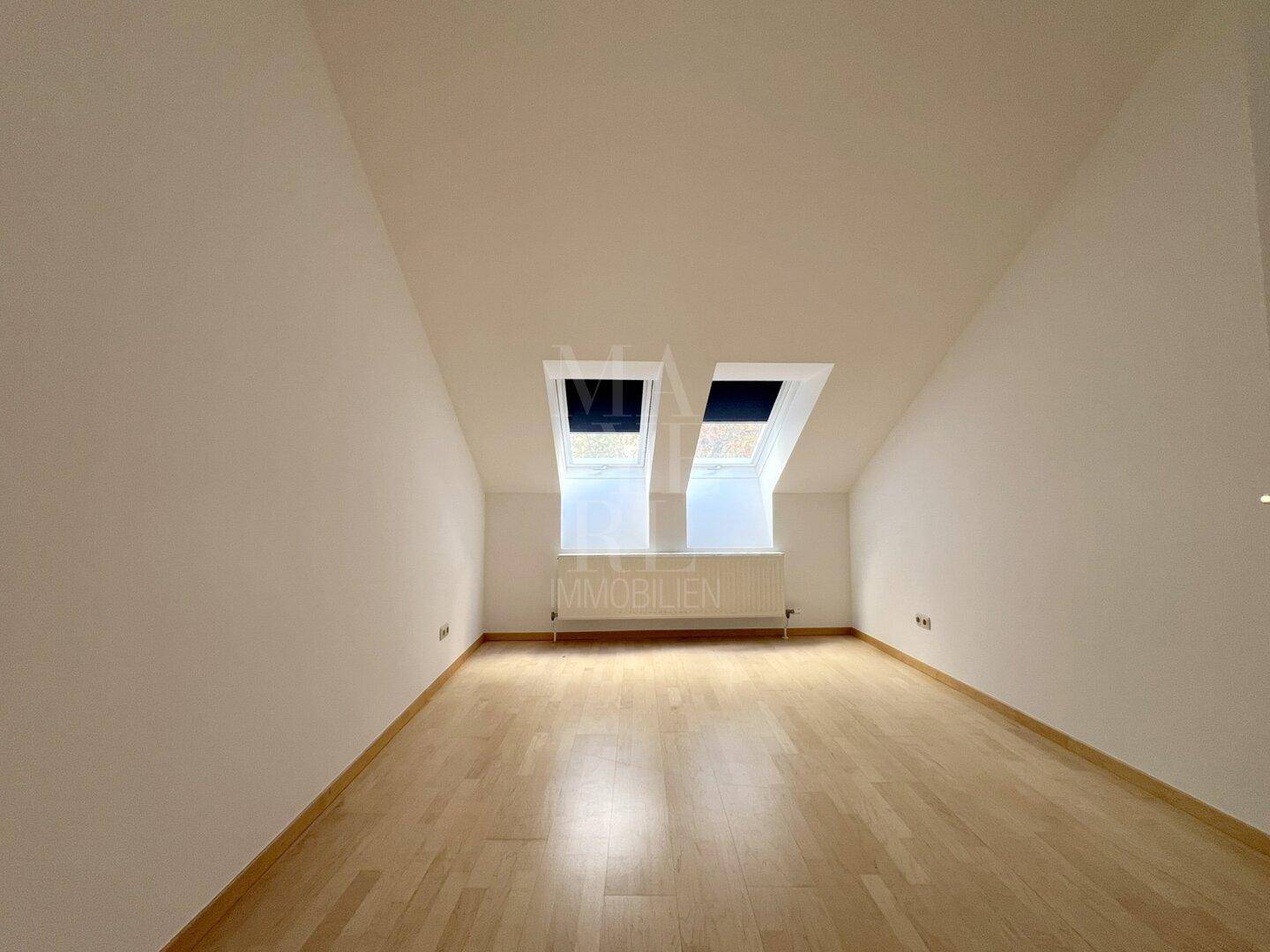 Helles Dachzimmer mit schrägen Decken, Holzboden und zwei Dachfenstern für viel natürliches Licht.