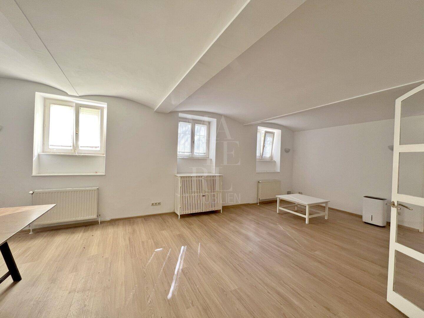 Geräumiges Zimmer mit hellem Holzboden, Gewölbedecke und mehreren Fenstern, die viel Licht spenden.