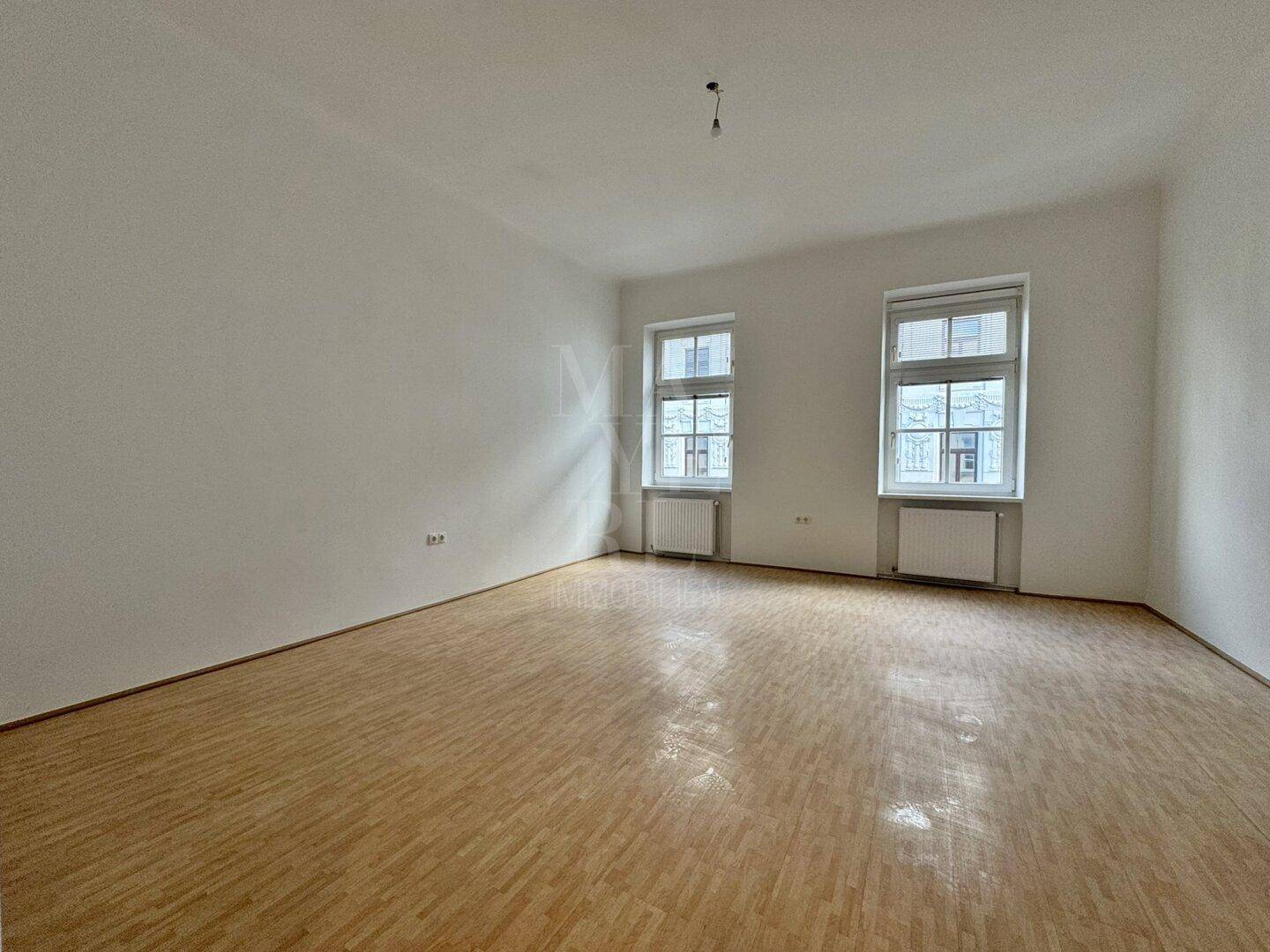 Geräumiges, unmöbliertes Zimmer mit Holzboden und Blick nach draußen durch zwei Fenster.