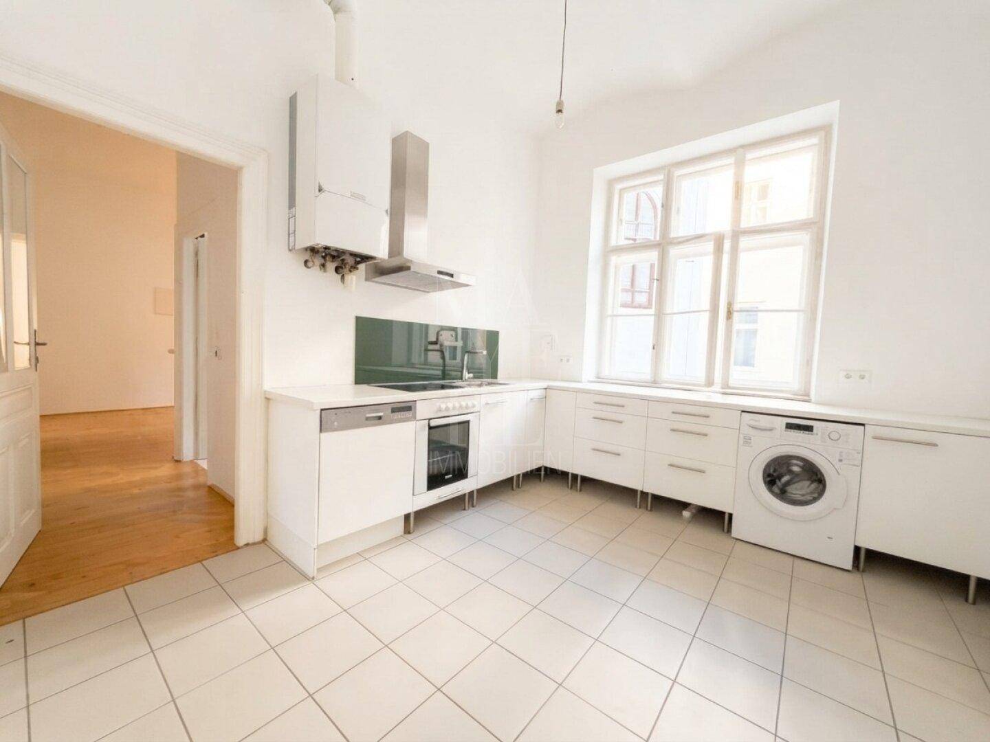 Helle Küche mit weißen Einbauschränken, Fliesenboden und großem Fenster, ausgestattet mit modernen Geräten und Waschmaschine.