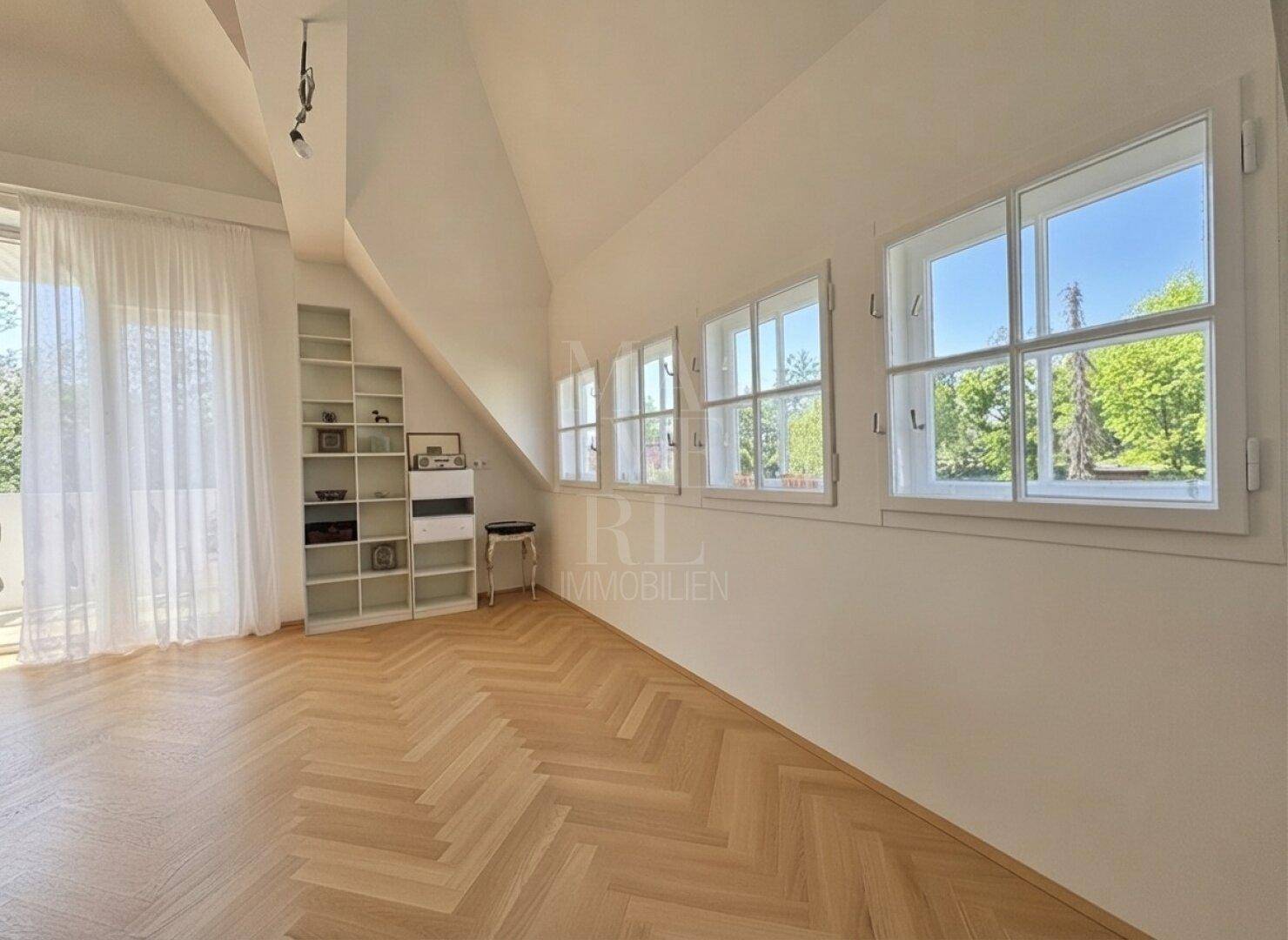 Heller Raum mit Fischgrätparkett, Schrägdach und einer Reihe von Fenstern mit Grünblick.
