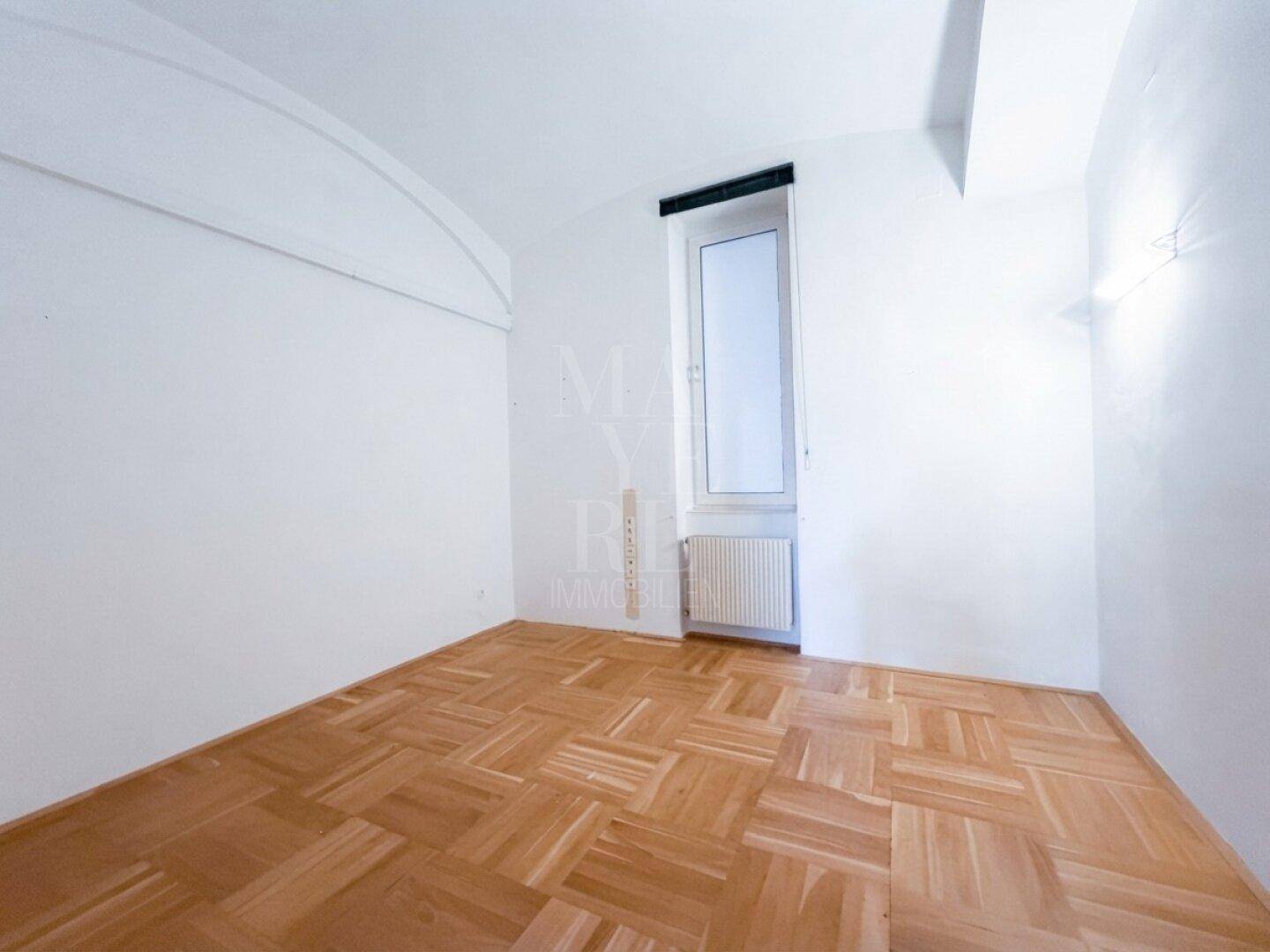 Helles Zimmer mit Parkettboden und einem hohen, schmalen Fenster, das den Raum erhellt.
