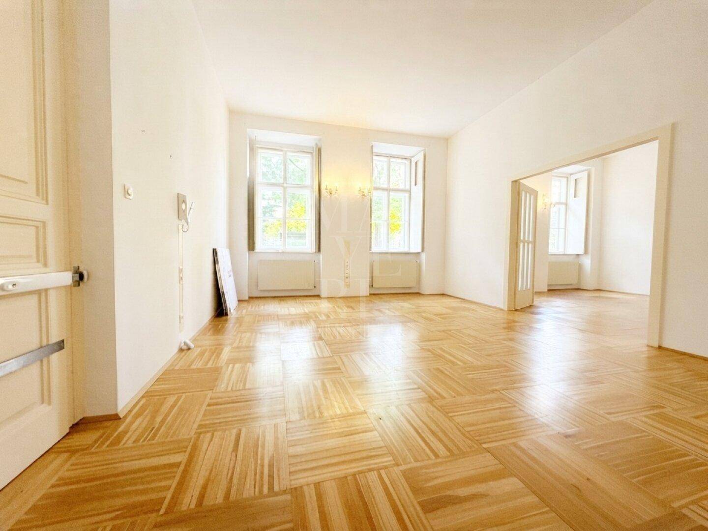 Heller Wohnbereich mit klassischem Parkettboden und hohen Fenstern, die den Raum mit natürlichem Licht füllen.