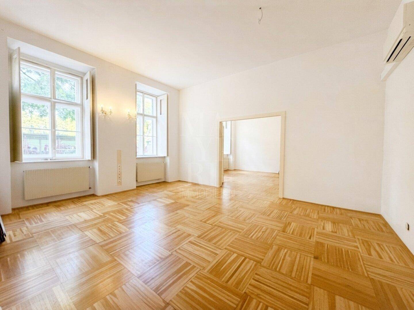 Großzügiger Wohnraum mit elegantem Parkett und zwei hohen Fenstern, die einen Blick nach draußen ermöglichen.