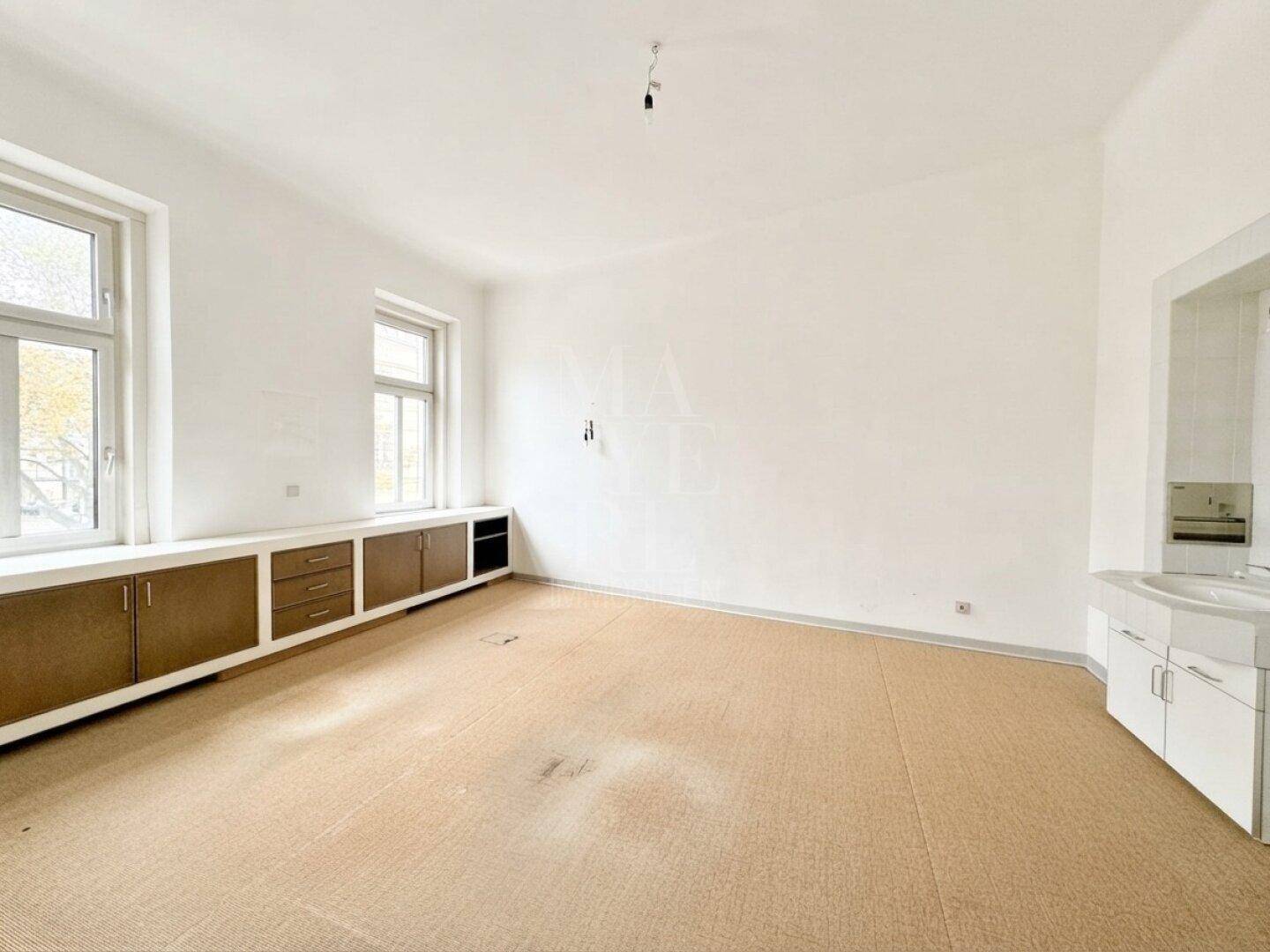 Heller Raum mit Teppichboden, Einbauschränken unter den Fenstern und einem Waschbeckenbereich an der Seite.