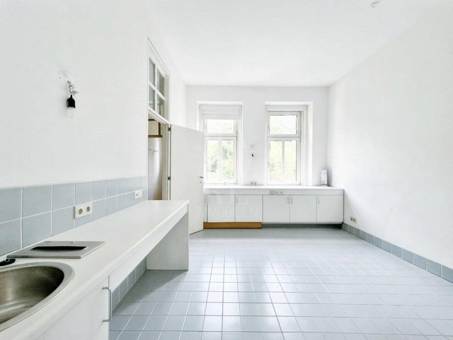 Helle Küche mit hellblauen Fliesen, einer langen Arbeitsplatte mit Spüle und Kochfeld sowie zwei Fenstern.