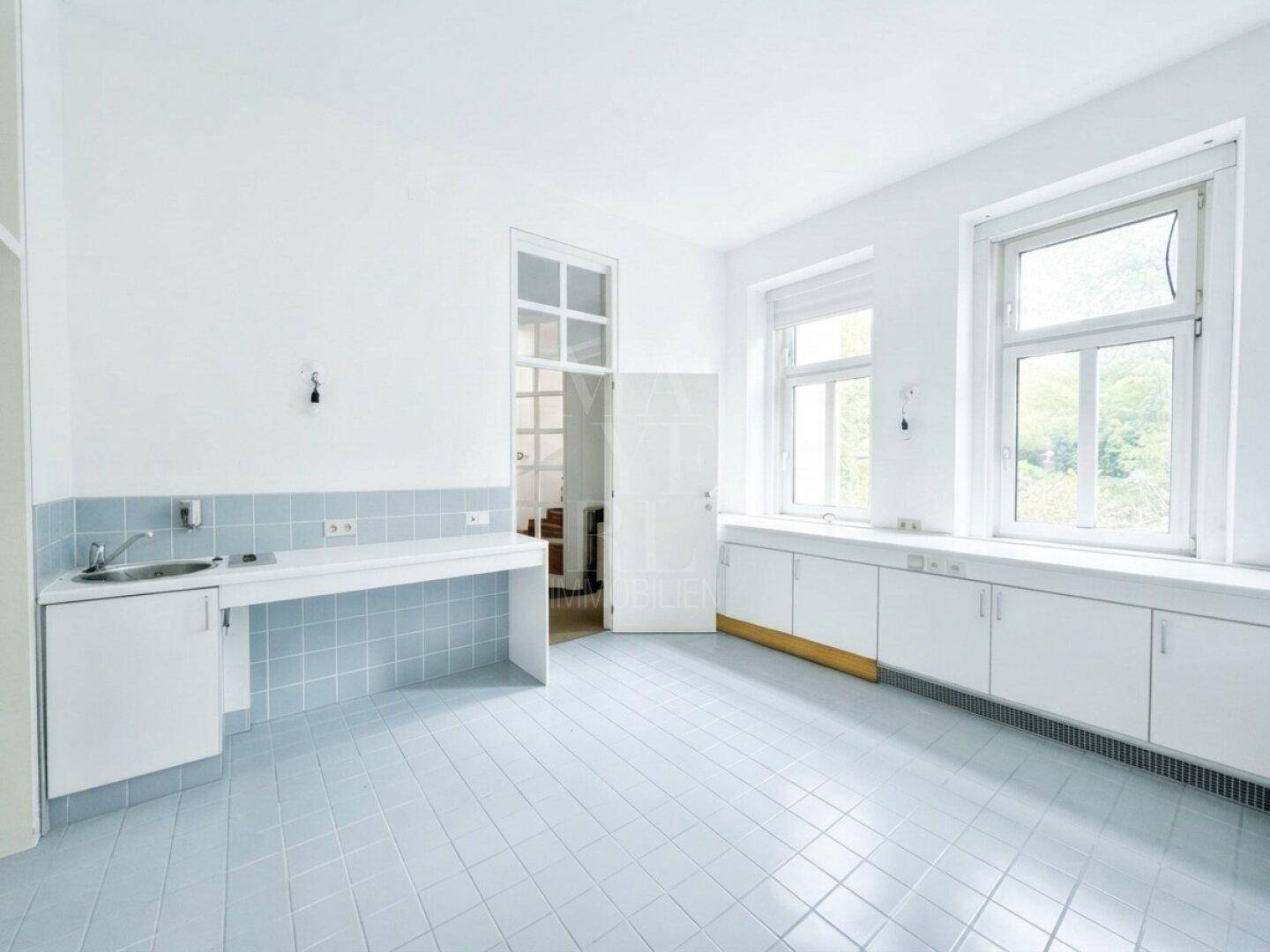 Geräumige Küche mit hellblauen Bodenfliesen, weißen Schränken unter den Fenstern und einer Arbeitsplatte mit Spülbecken.