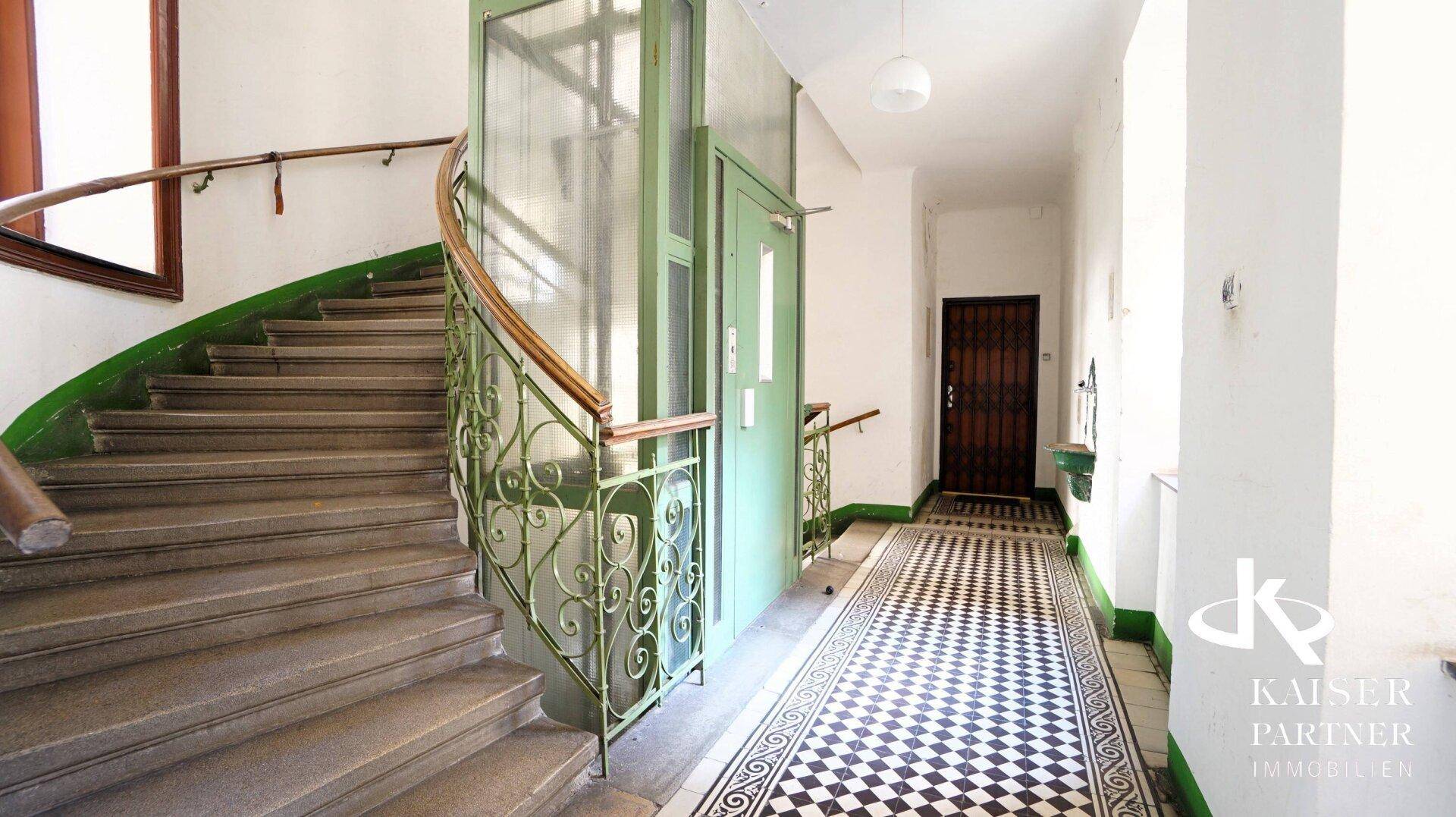 Historisches Treppenhaus mit geschwungener Treppe, Schachbrettfliesen und einem grünen Aufzug.