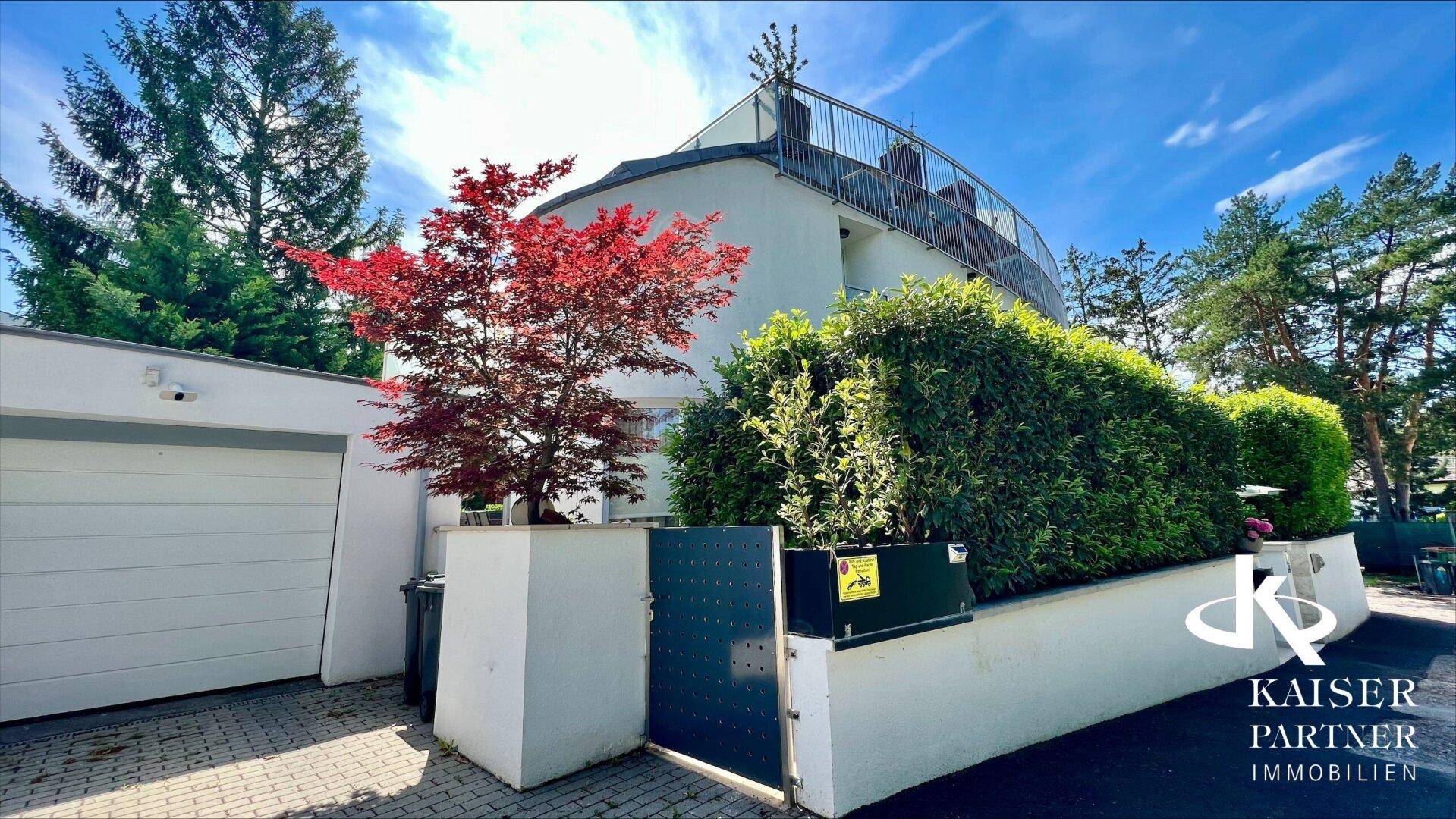 Eingangsbereich des modernen Gebäudes mit Garagentor, Hecke und einem auffälligen roten Baum.