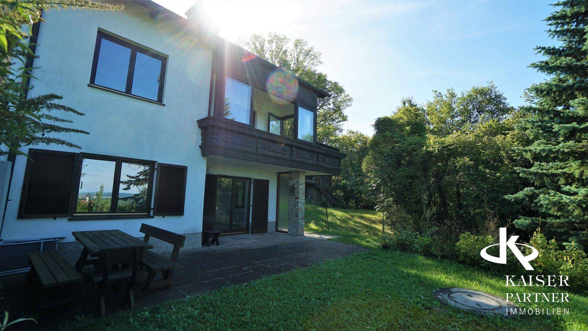 Hausfassade mit Terrasse und Gartenbereich, umgeben von Bäumen unter strahlendem Sonnenschein.