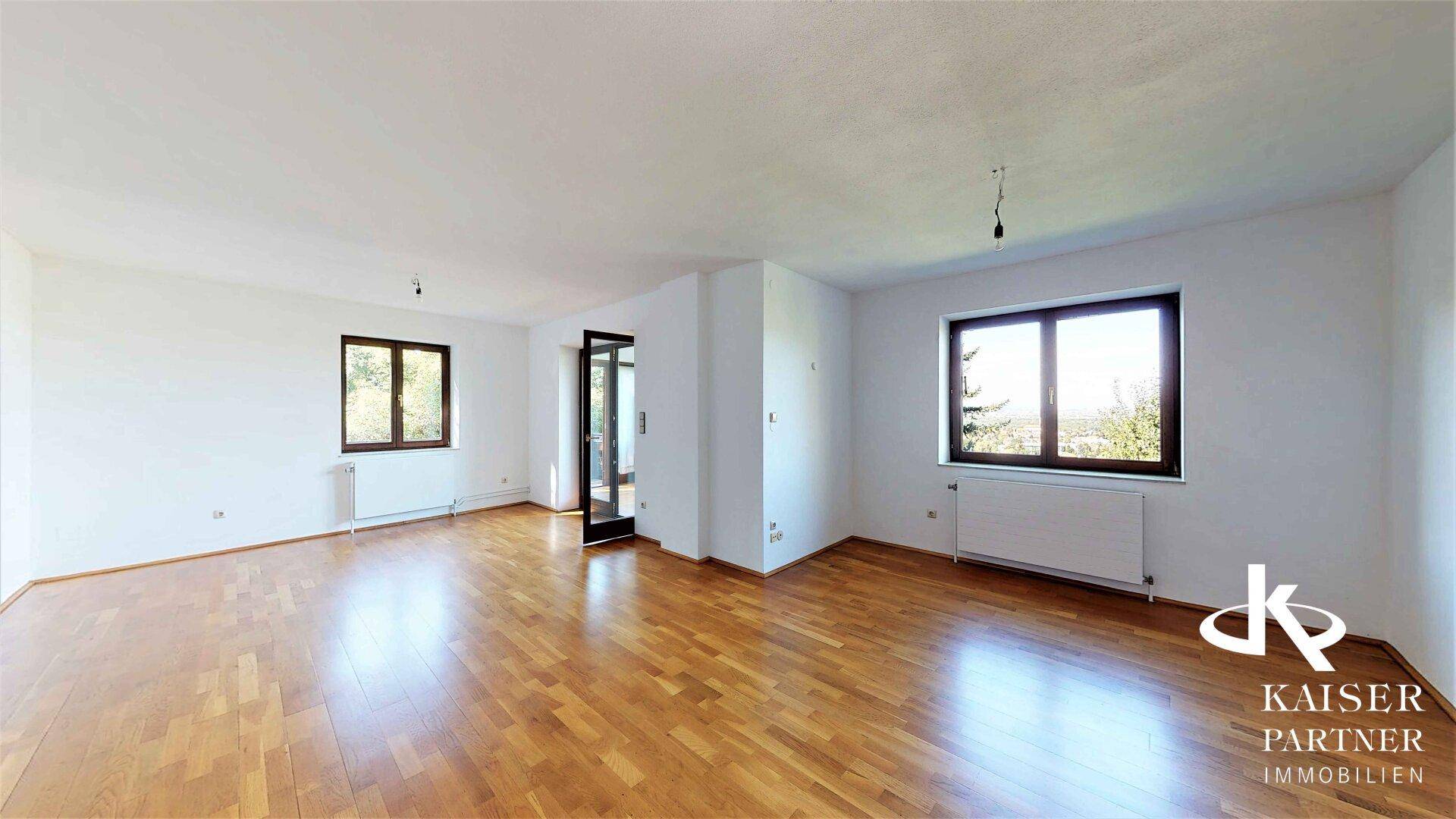 Geräumiges Wohnzimmer mit hellem Parkettboden und großen Fenstern, die viel Tageslicht hereinlassen.