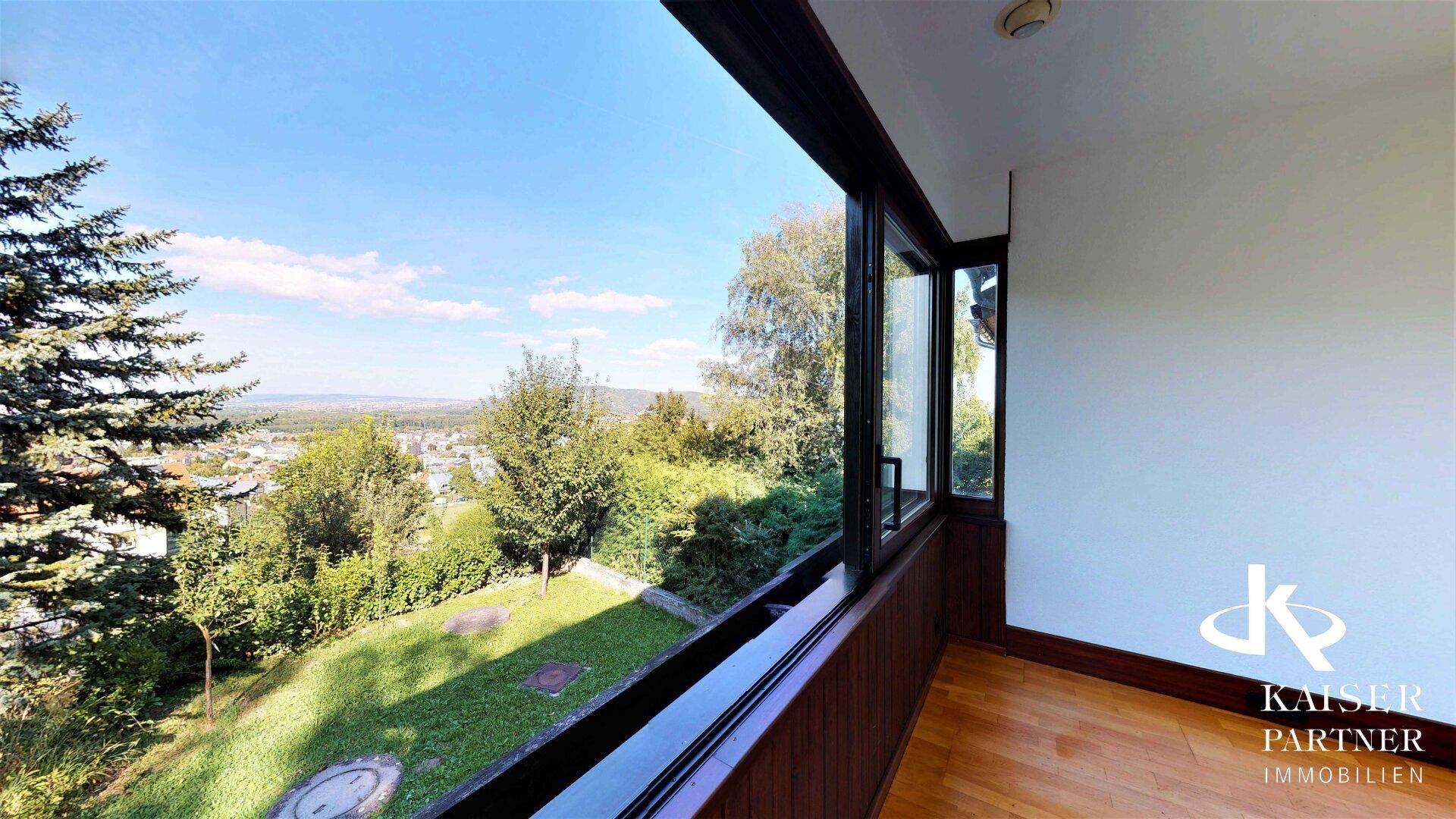 Heller Raum mit breiten Fenstern, die einen weiten Blick über den Garten und die Stadt bieten.