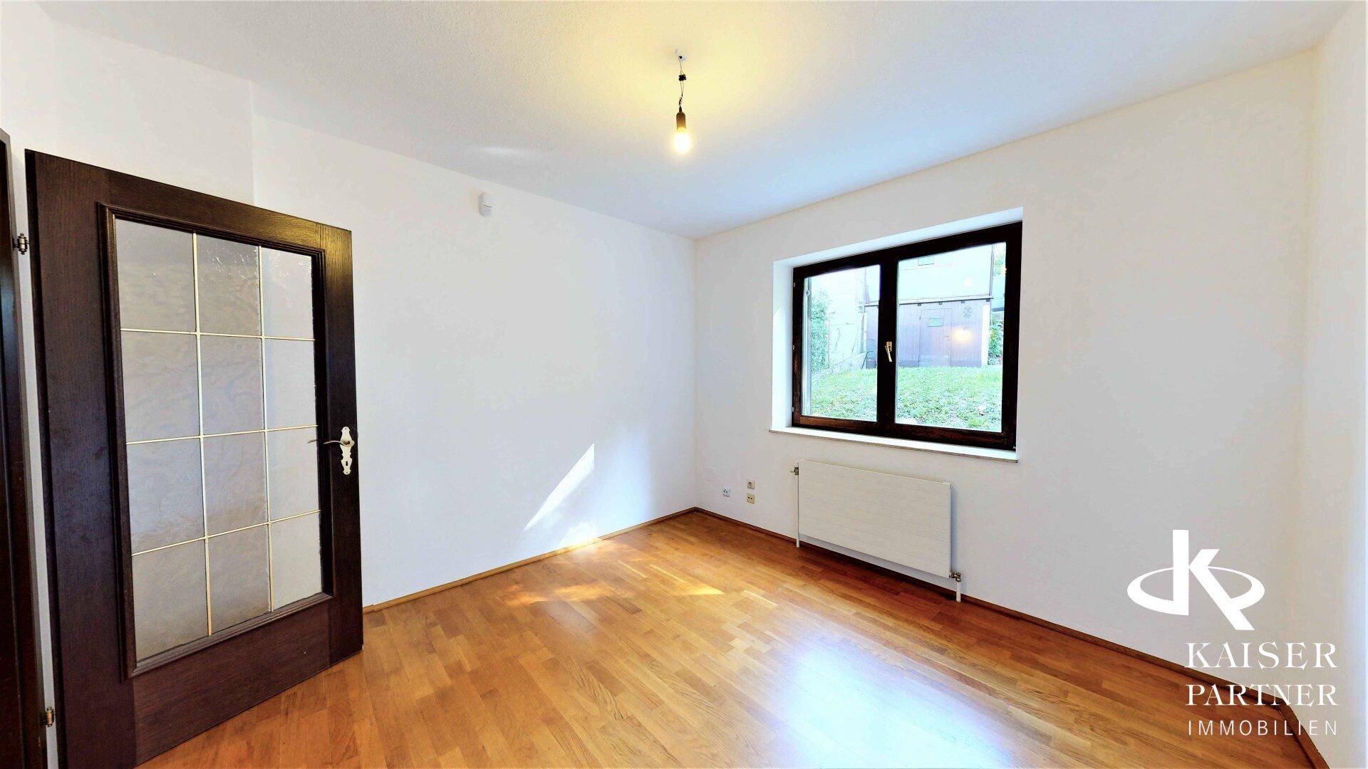 Helles Schlafzimmer mit Parkettboden und einem Fenster, das den Blick ins Grüne freigibt.