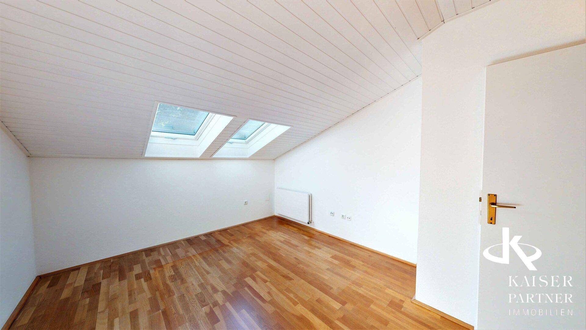 Dachzimmer mit schräger Decke, Parkettboden und zwei Dachfenstern, die viel Licht spenden.