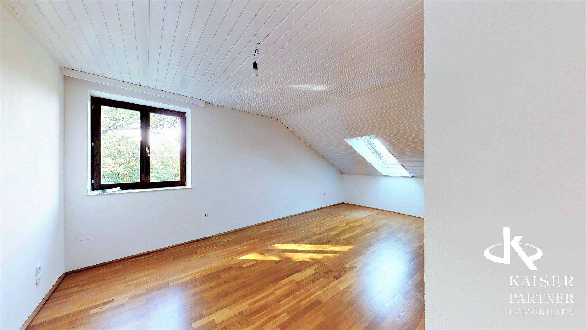Helles Dachzimmer mit Holzdecken, Parkettboden und einem Fenster mit Blick ins Grüne.