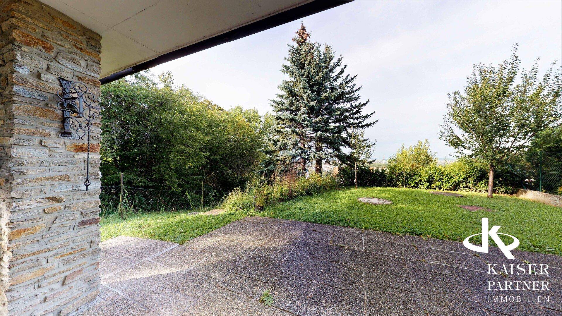 Gepflasterte Terrasse mit Natursteinwand und Blick auf den grünen Garten und die Bäume.