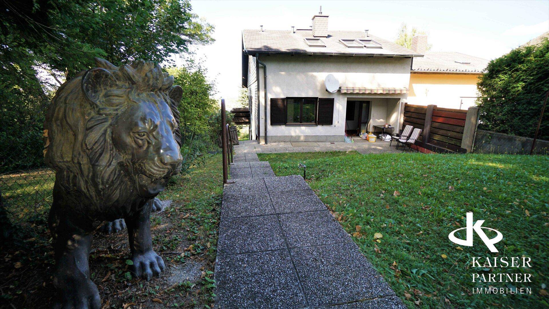 Grüner Gartenbereich mit gepflastertem Weg, der zum Haus führt, und einer dekorativen Löwenstatue.
