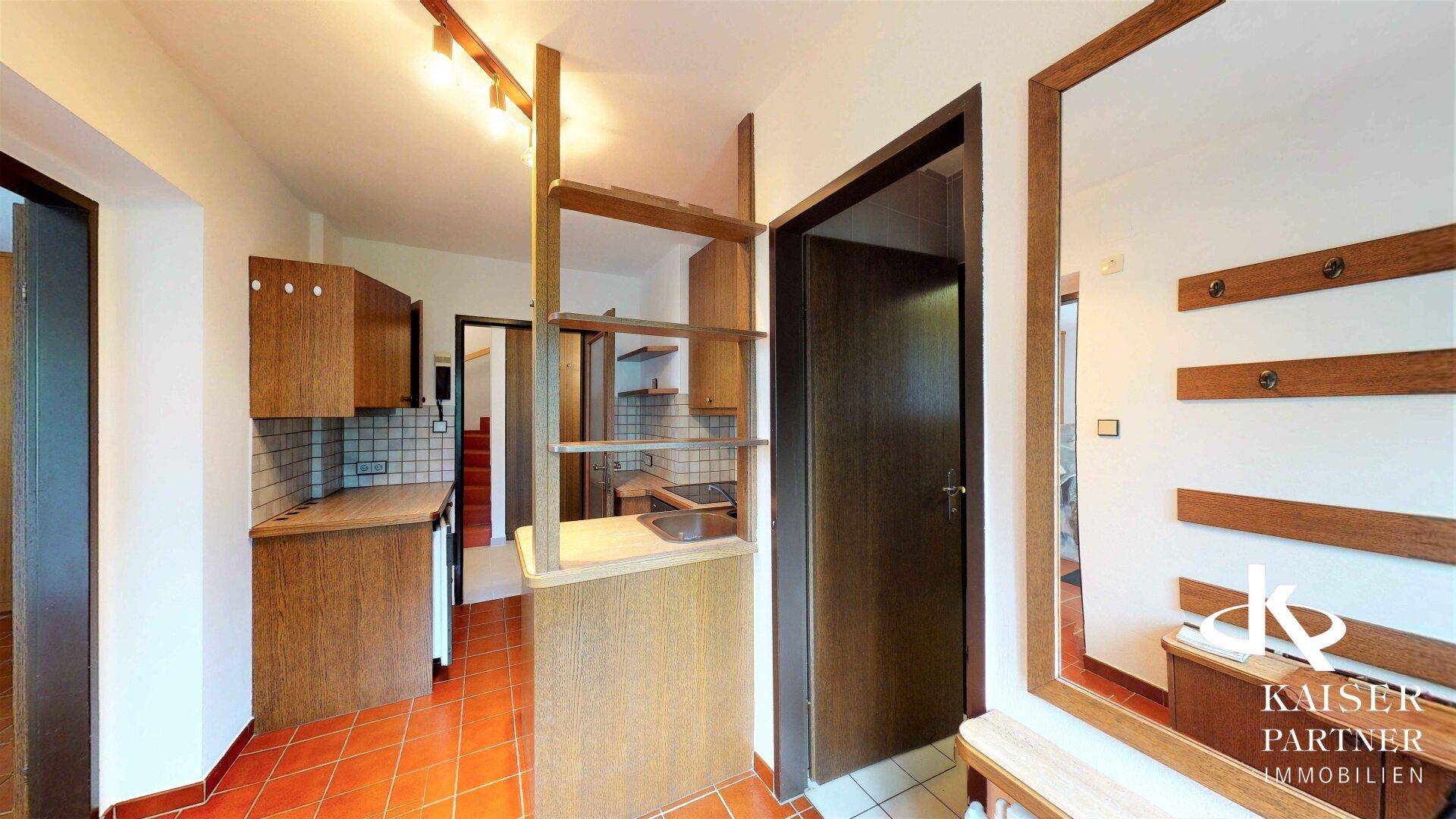 Funktionaler Eingangsbereich mit integrierter Küchenzeile, Holzschränken und Terrakotta-Fliesenboden.