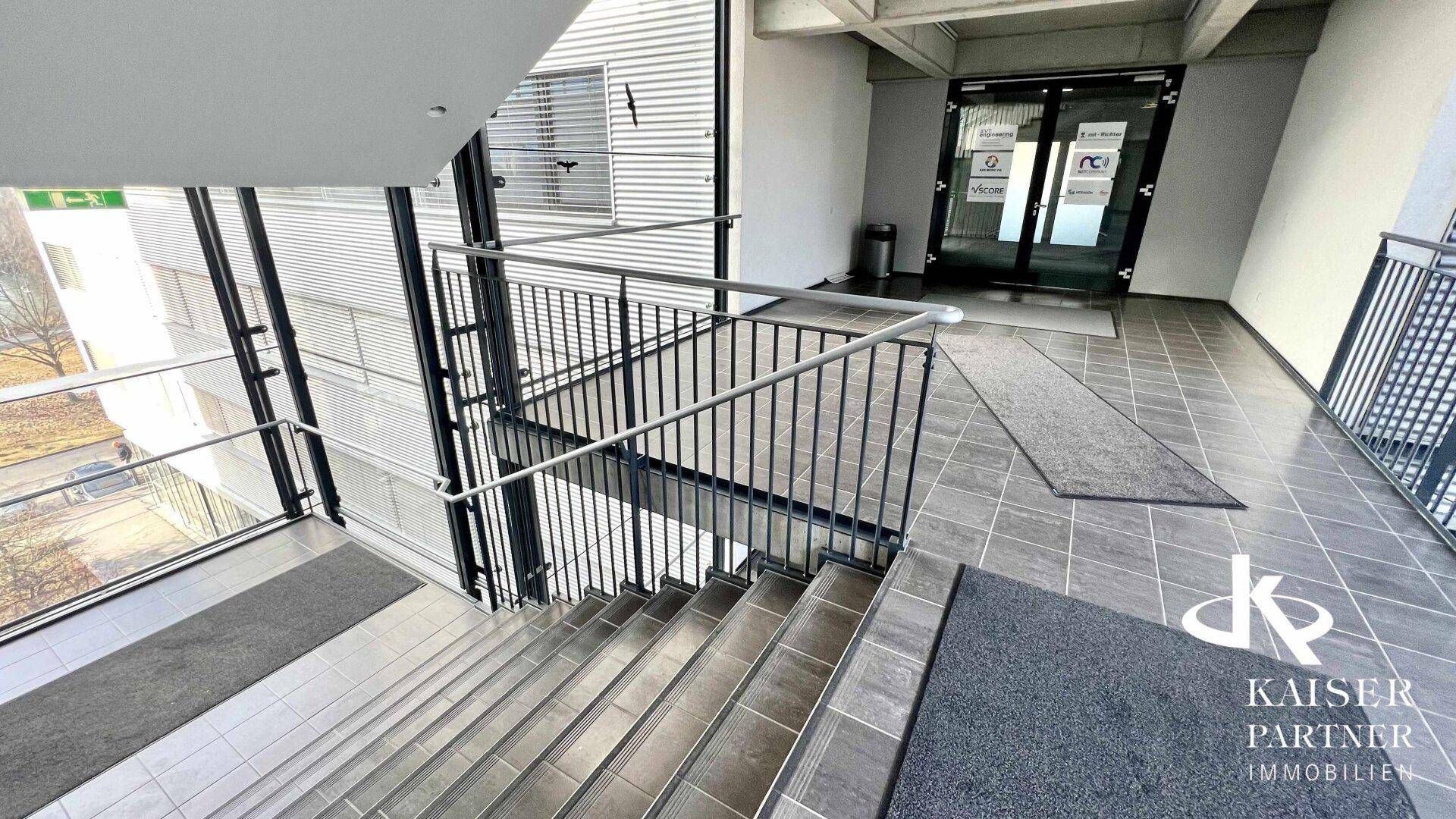 Moderner Treppenbereich mit Fliesenboden und Metallgeländer, führt zum Eingang mit Glastüren.