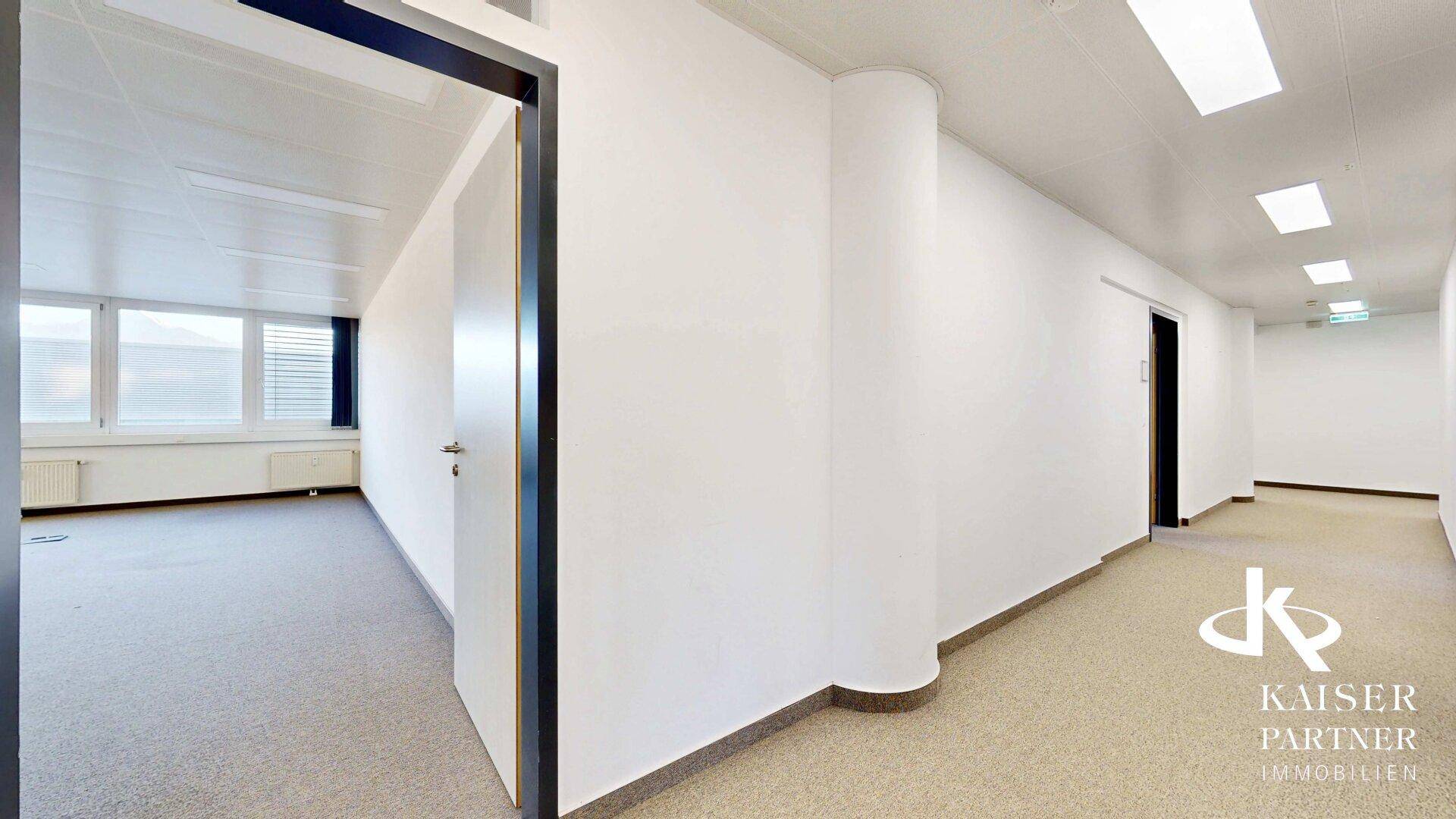 Heller Bürogang mit Teppichboden und Zugang zu einem leeren Büroraum mit Fenstern.