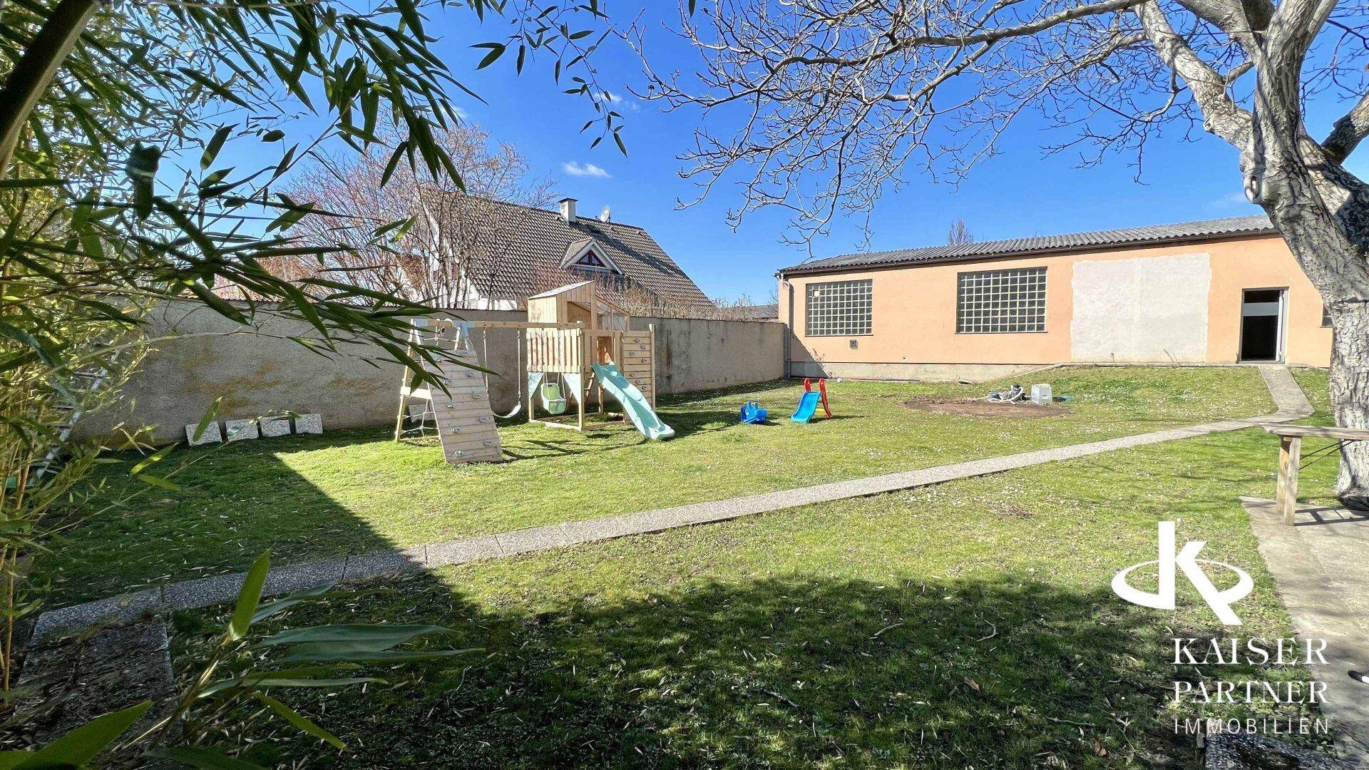 Großer Gartenbereich mit Rasenfläche, Kinderspielplatz und Nebengebäude unter strahlendem Himmel.